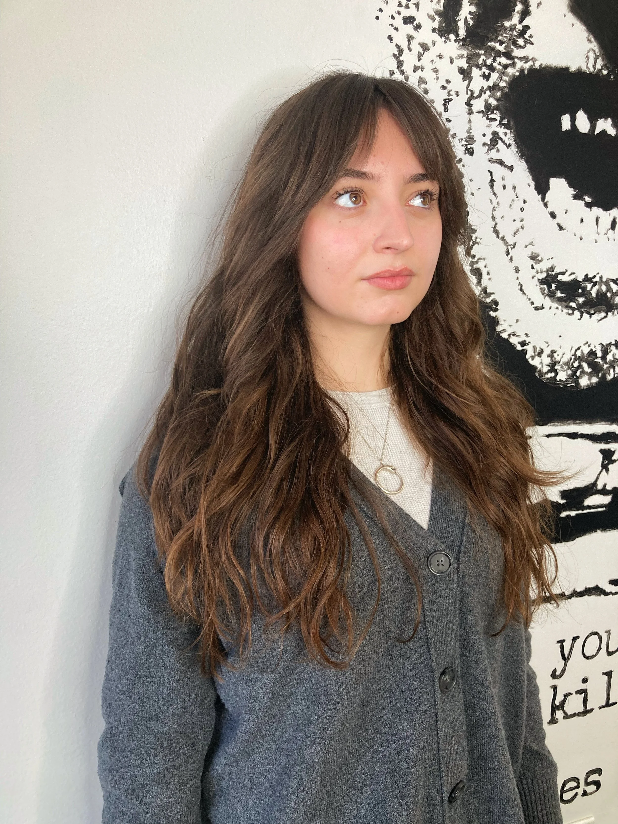 Young woman with long wavy brown hair wearing a gray cardigan, white shirt, and a gold necklace, standing against a white wall with a black and white artwork behind her.