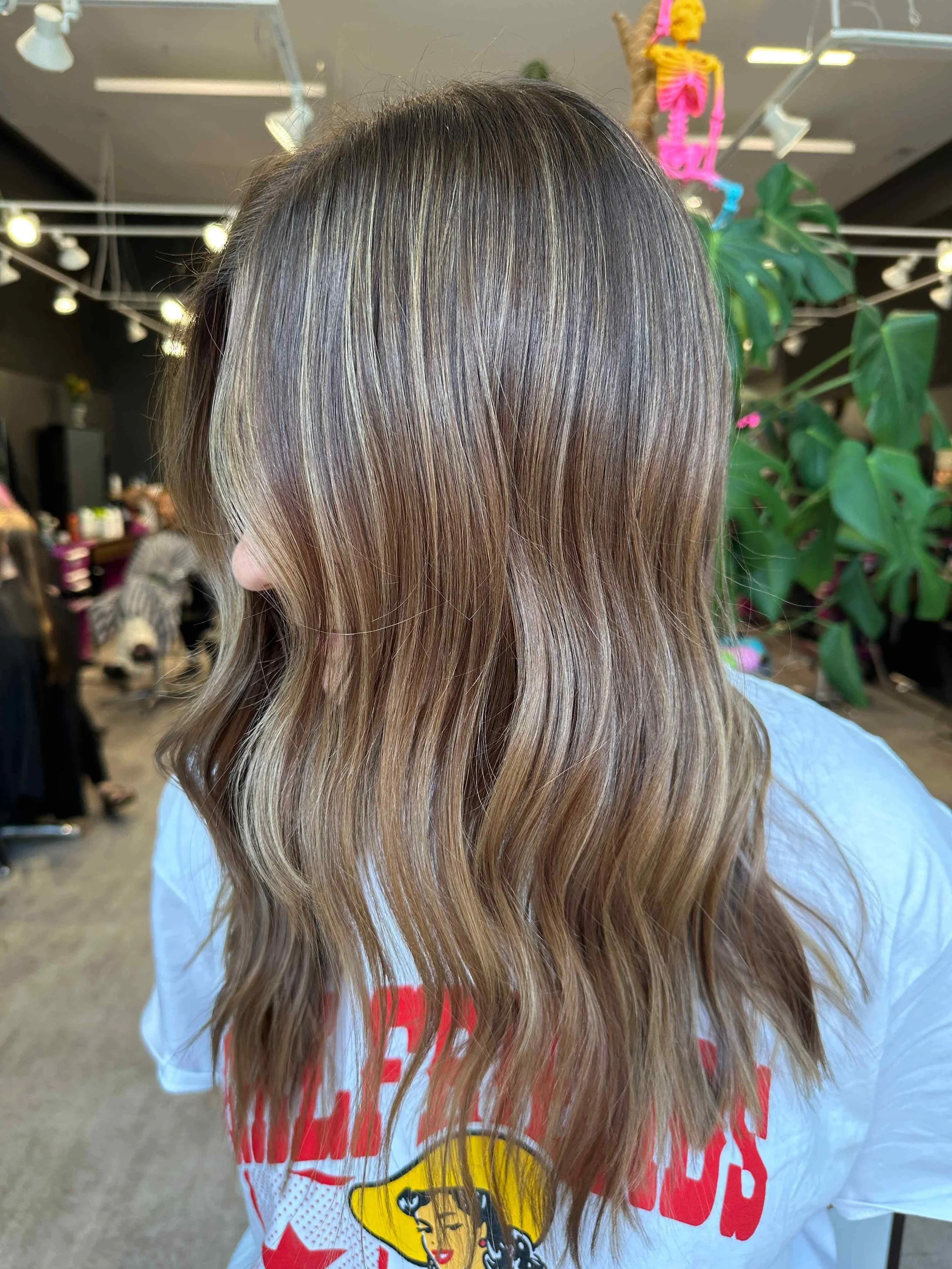A person with long, wavy hair dyed in a balayage style, blending dark roots with light brown and blonde highlights, standing in a hair salon with salon lighting, plants, and other people in the background.