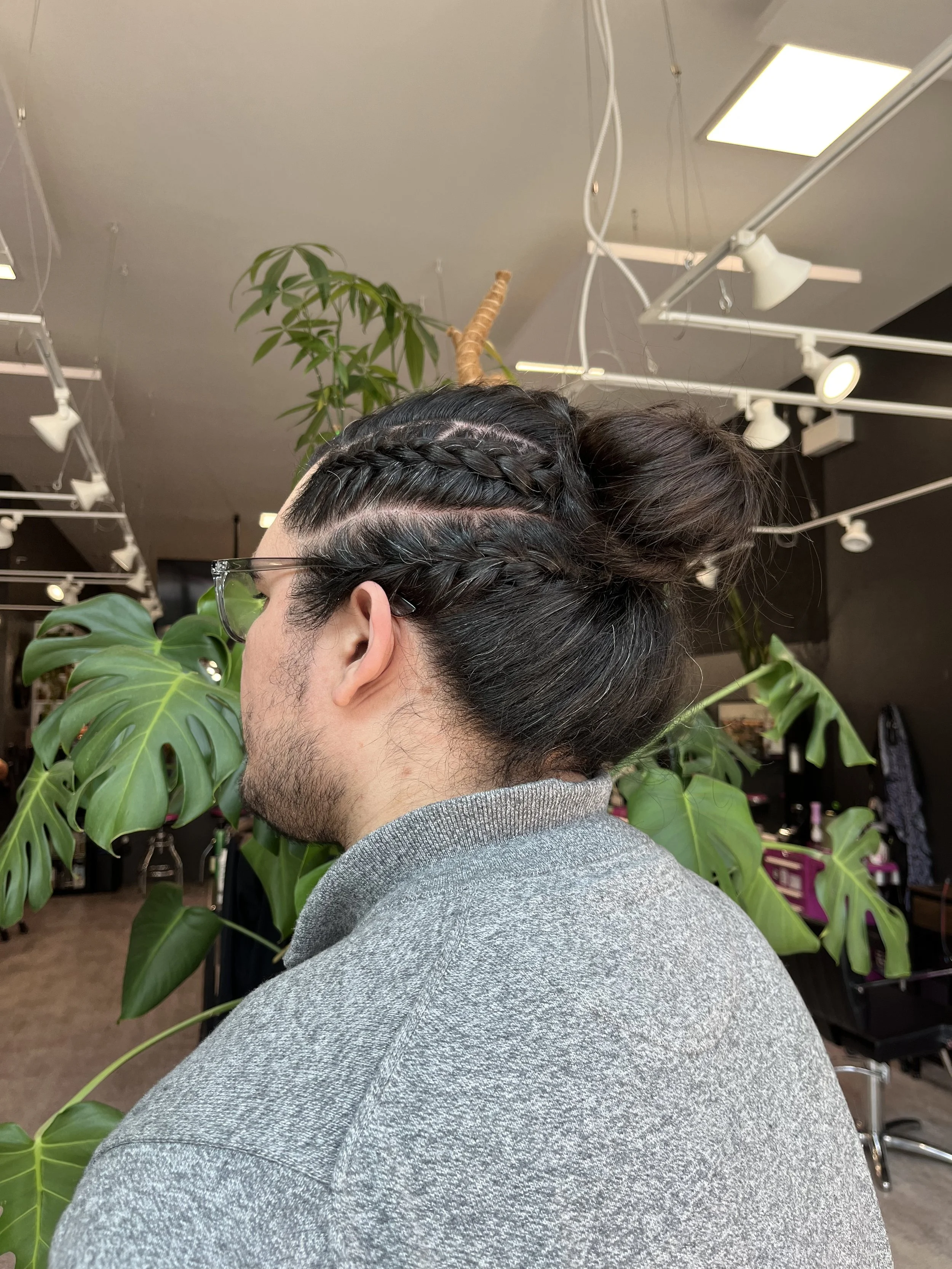A person with dark brown hair styled in multiple braids and a bun, wearing glasses and a gray hoodie, standing amidst green Monstera plants in an indoor setting.