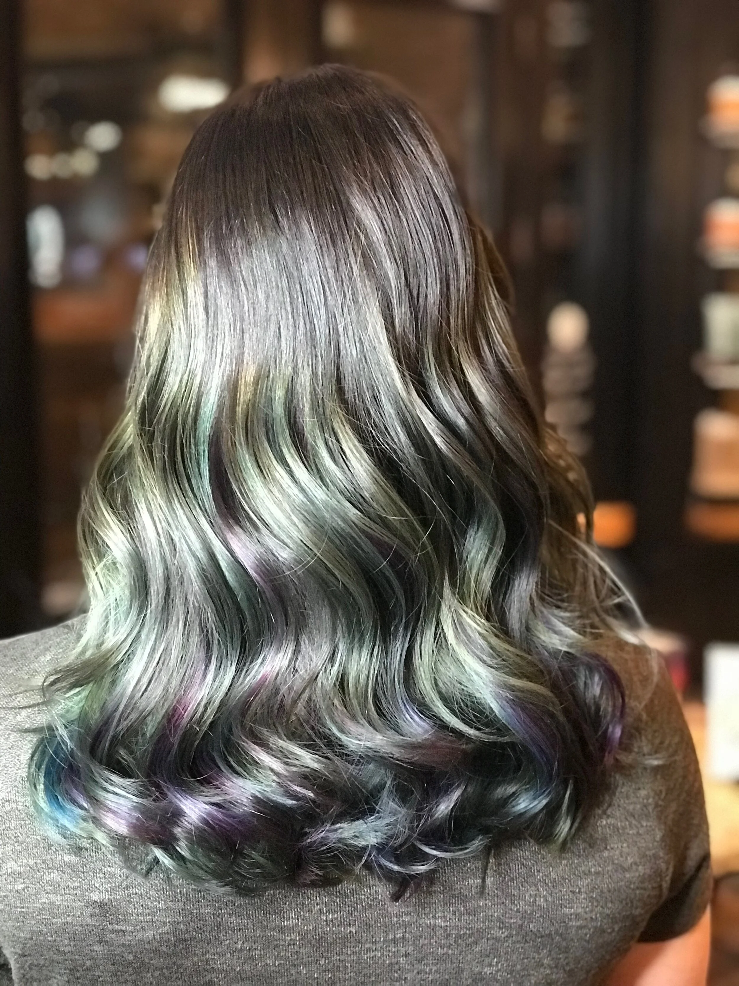 Back of a person's head with long, wavy hair styled with subtle colorful highlights including green, purple, and blue.