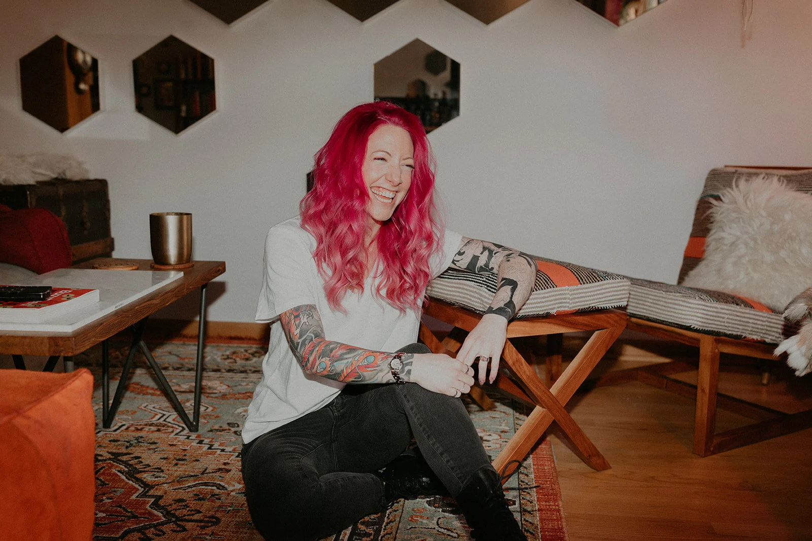 A woman with pink hair and colorful tattoos sitting on the floor in a living room, smiling and laughing.