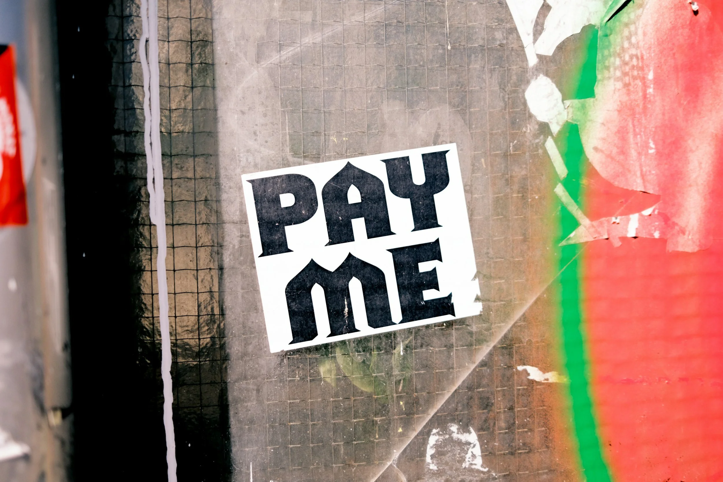 A sticker with the words 'Pay Me' in bold black letters on a white background, placed on a wire fence, with some graffiti and peeling paint visible nearby.