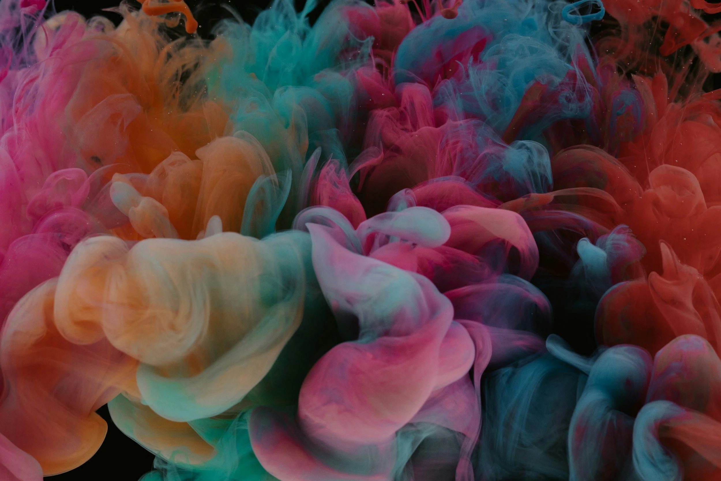 Colorful swirling clouds of ink in water close-up, featuring pink, orange, blue, and white hues against a black background.