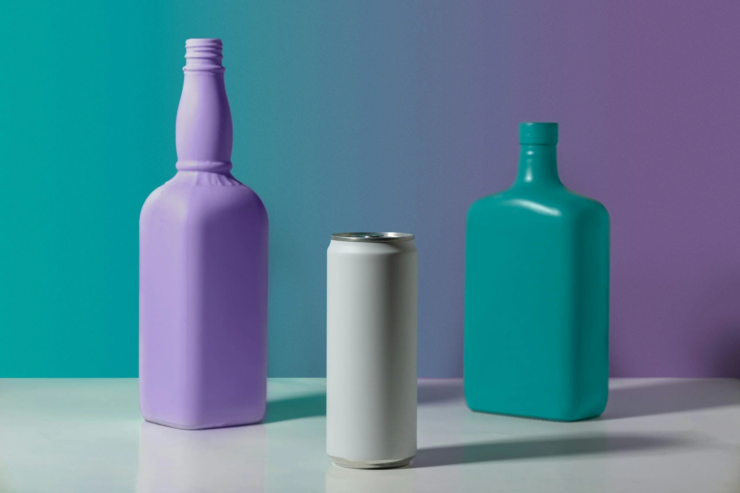 Two bottles, one painted purple and one teal, and a plain white can are displayed on a white surface against a gradient background shifting from teal to purple.