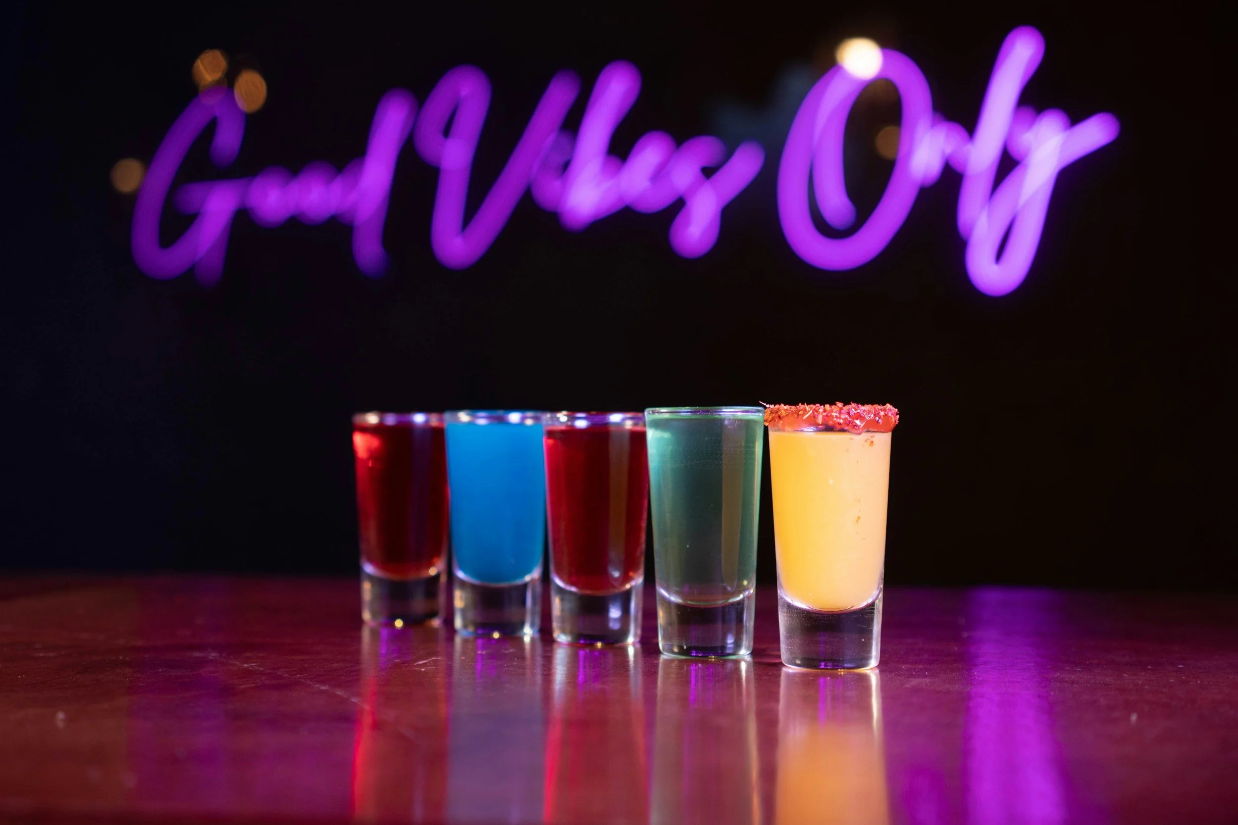 Five shot glasses filled with colorful drinks, lined up on a dark bar counter, with a neon sign that says 'Good Vibes Only' in purple lighting in the background.