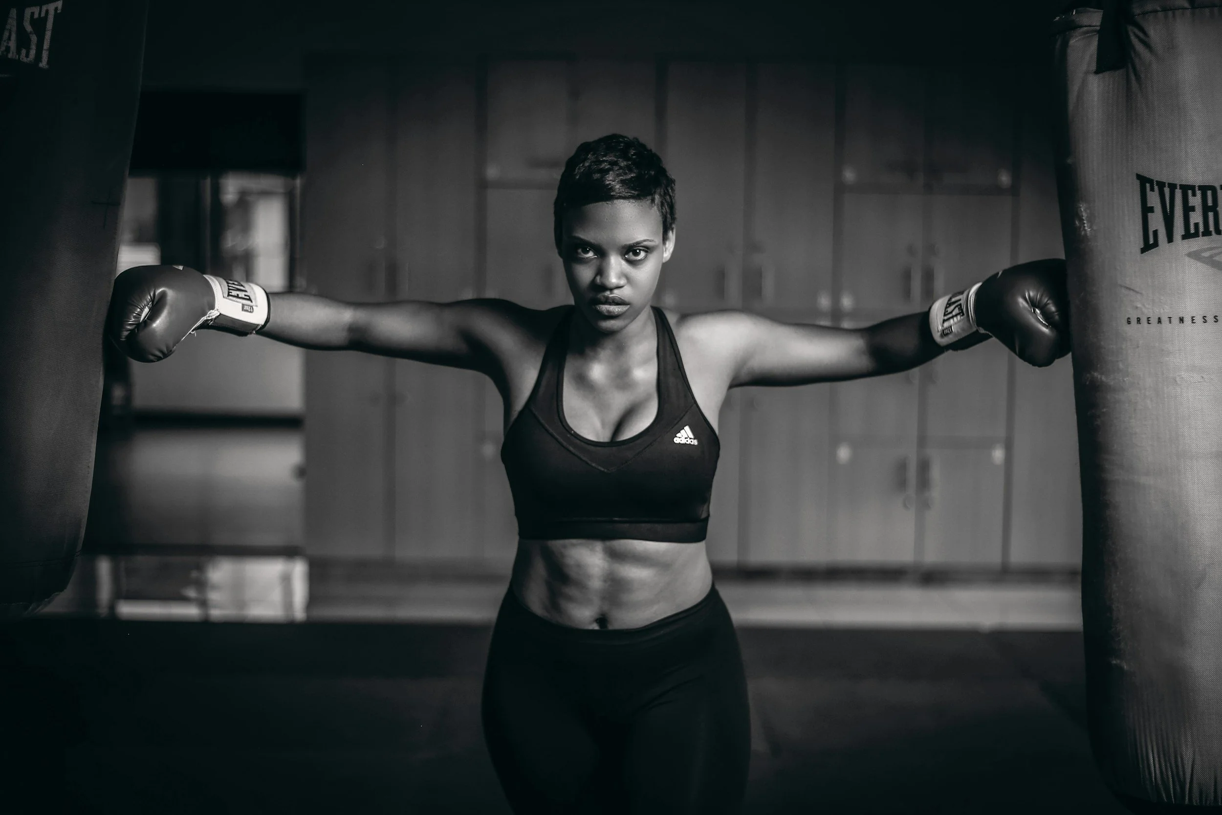 A fit woman in workout clothing boxing training in a gym, standing between punching bags with arms outstretched, looking intensely at the camera.
