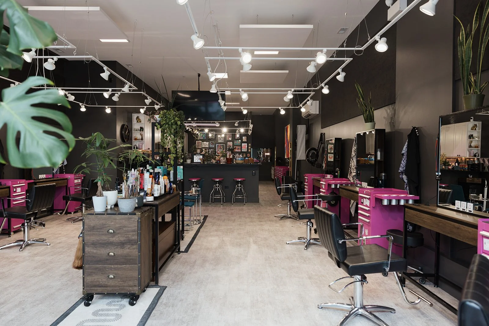 Interior of a modern hair salon with hair styling stations, mirrors, and black chairs with pink storage units. Hair products and tools are on the workstations, and plants are decorating the space.