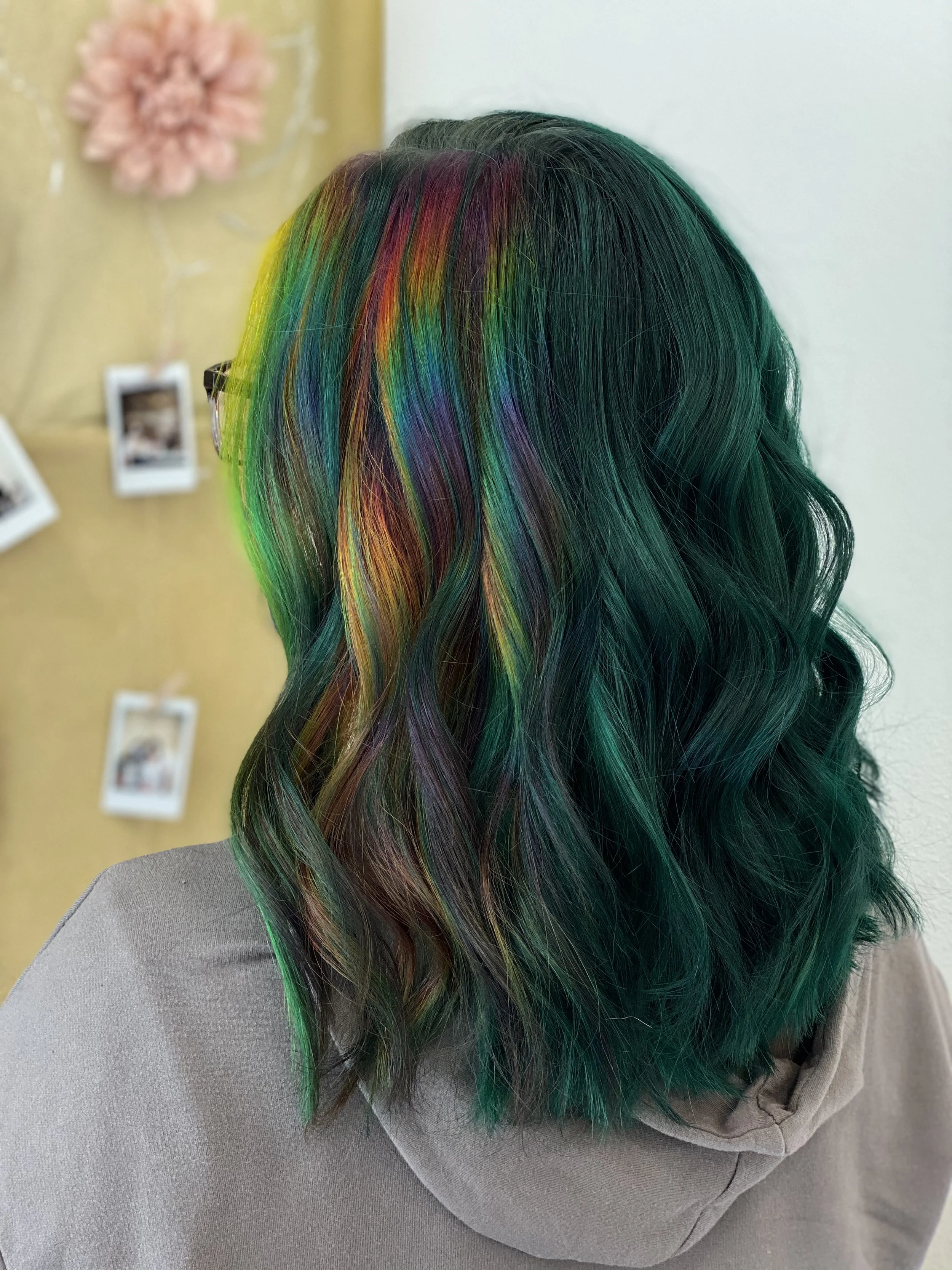 Back of a person's head with shoulder-length wavy hair dyed in multiple rainbow colors, including green, red, purple, yellow, and blue.