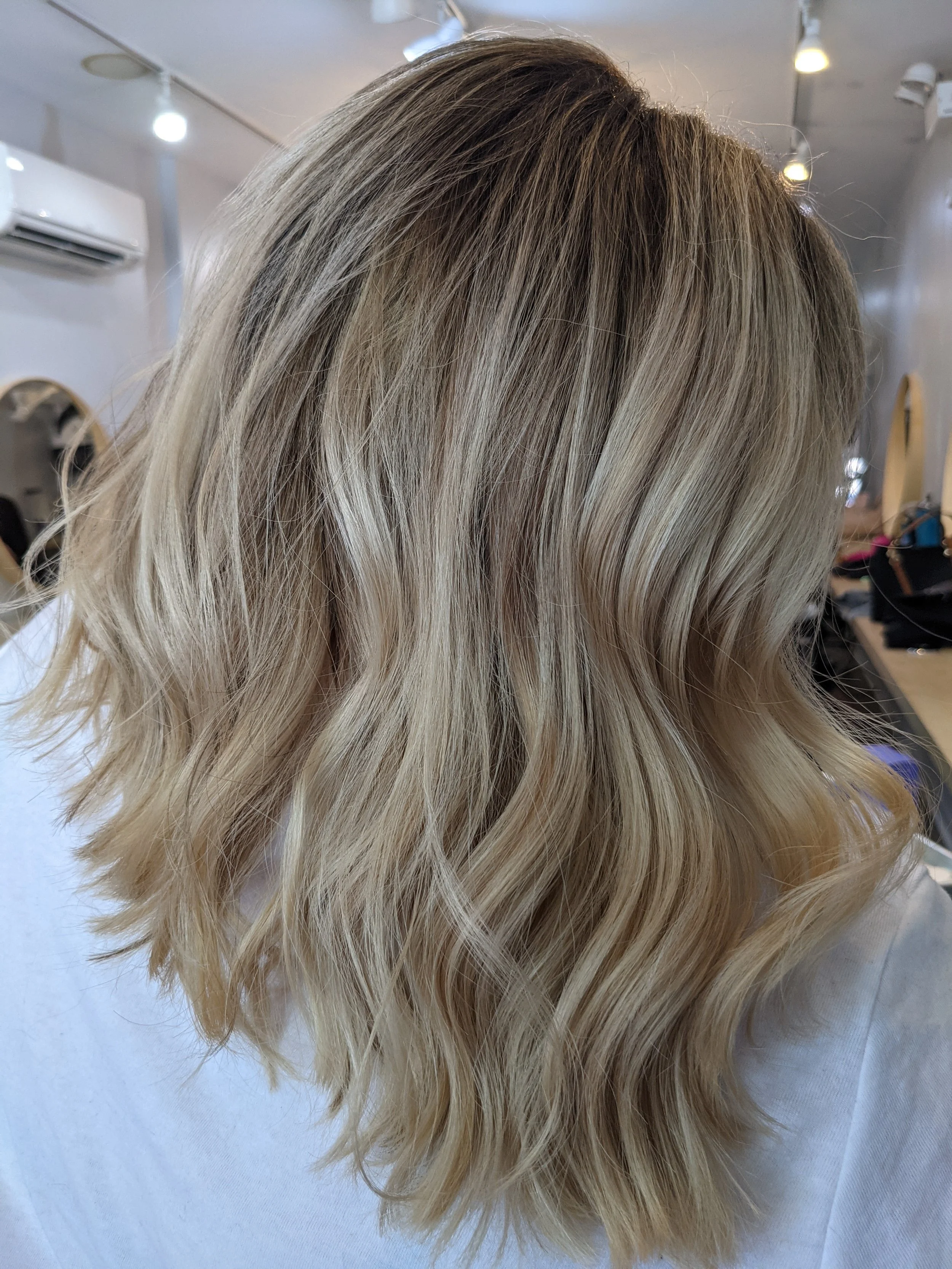 A close-up of a woman with shoulder-length, wavy blonde hair highlighted with darker roots, in a salon setting.