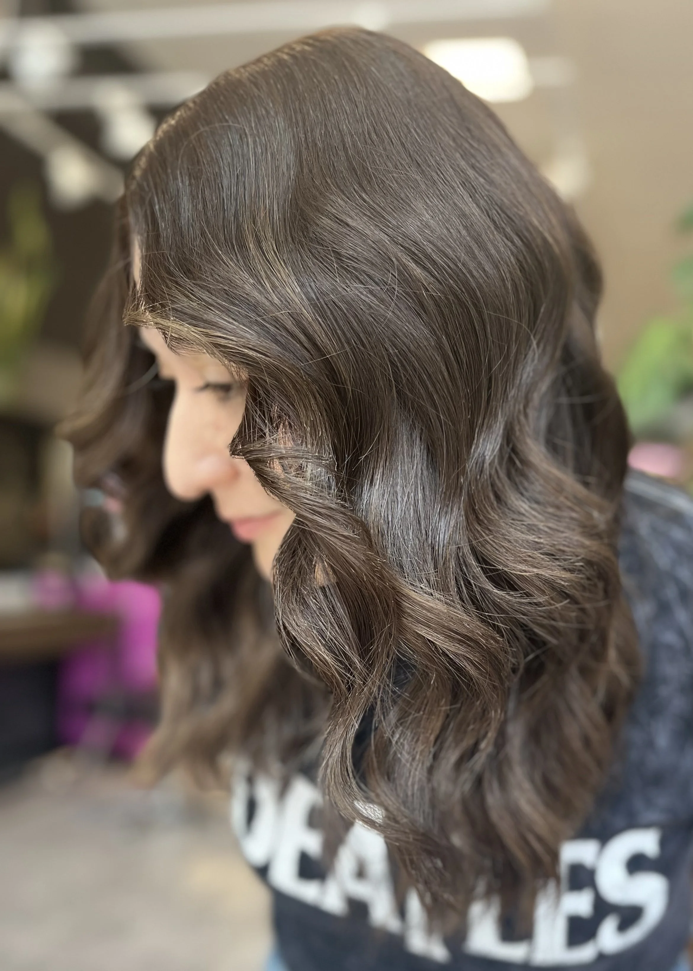 Close-up of a woman with shiny, wavy medium-length brown hair, looking down, in an indoor setting.