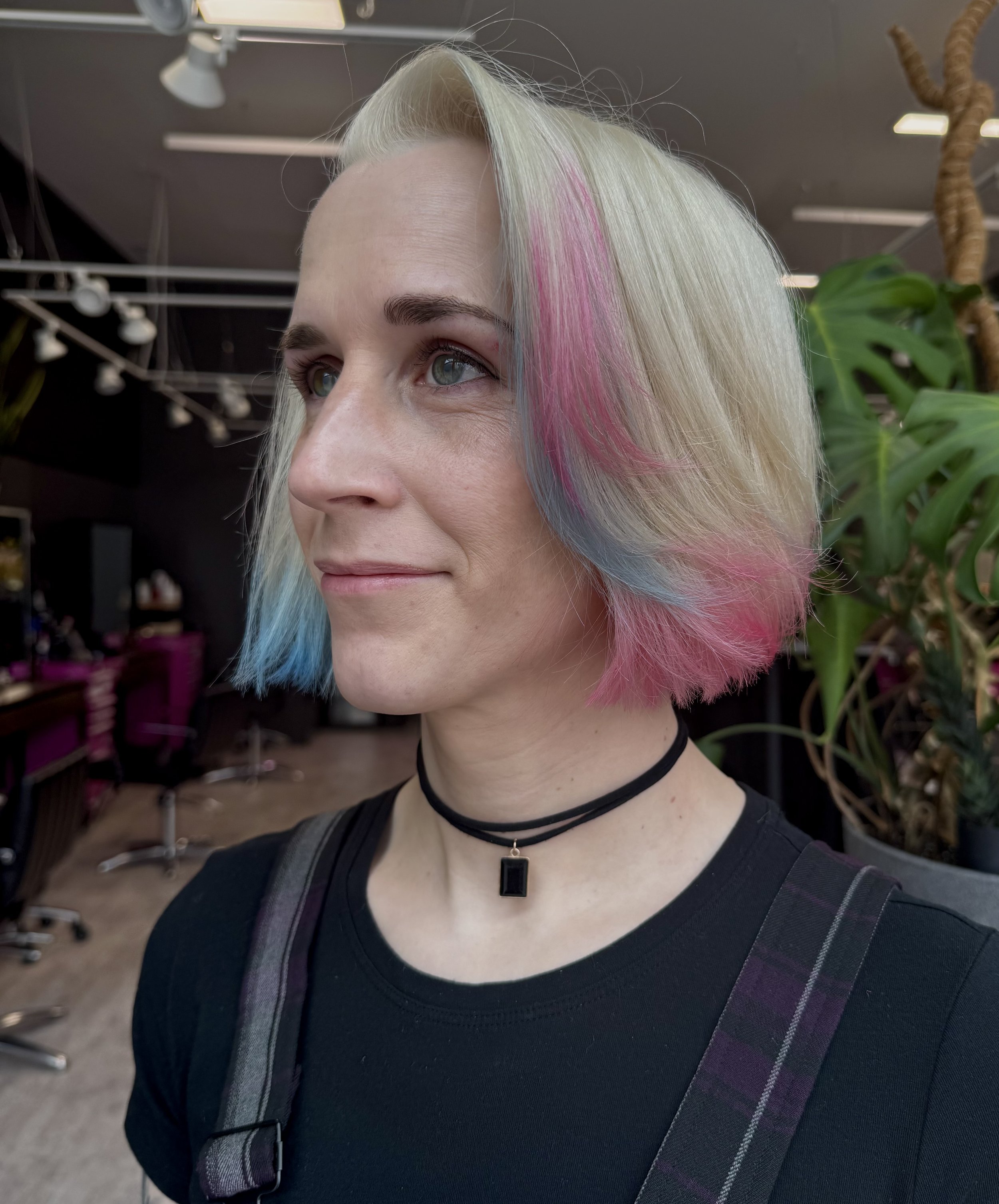 A woman with short, pastel-colored hair dyed pink, blue, and silver, wearing a black shirt, a black choker with a black pendant, and a dark-colored strap over her shoulder, standing in a salon with plants and hair styling chairs in the background.