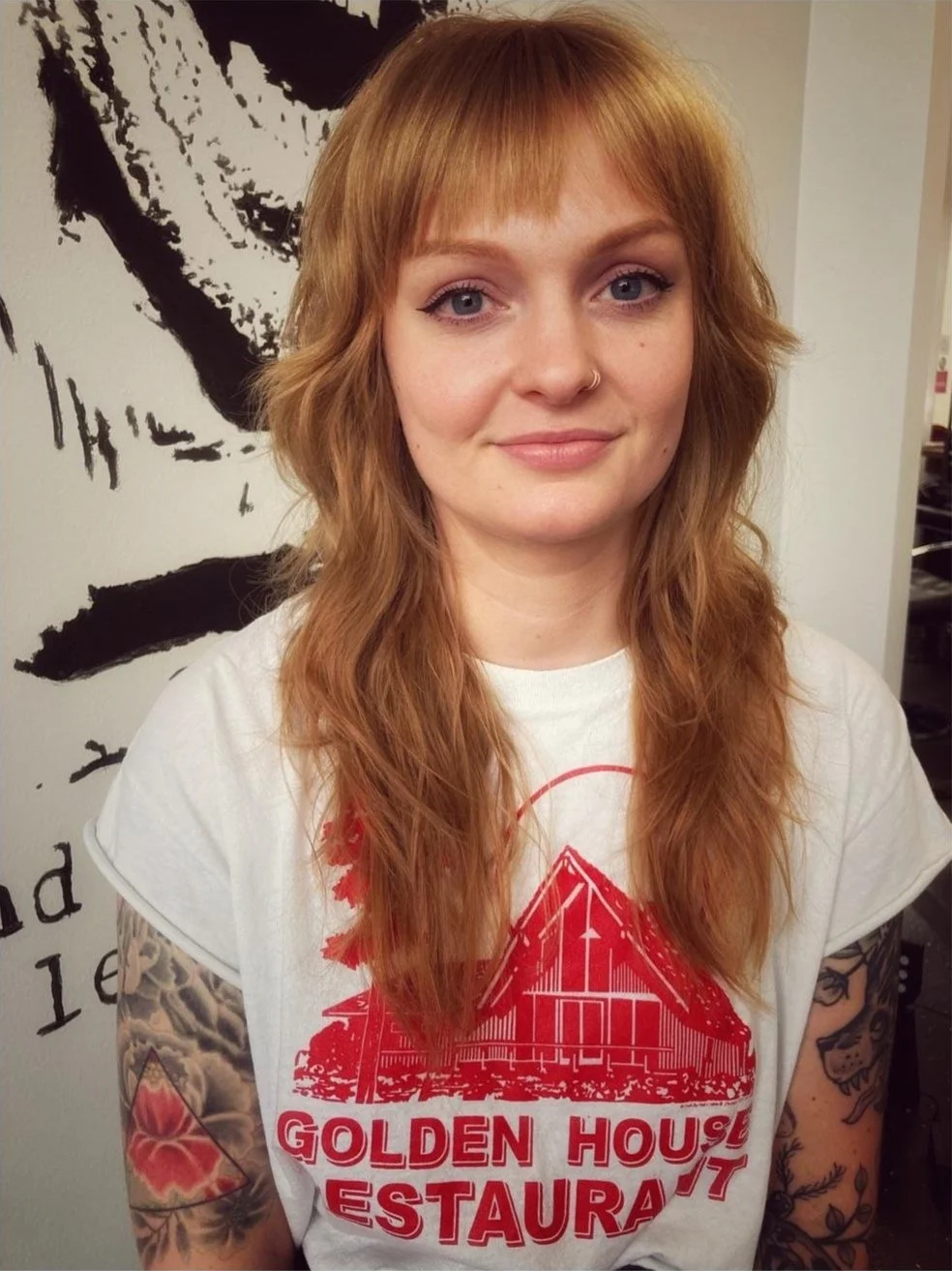A young woman with wavy red hair and a nose ring, wearing a white T-shirt with a red graphic and the words 'GOLDEN HOUSE RESTAURANT,' standing in front of a wall with black and white artwork.