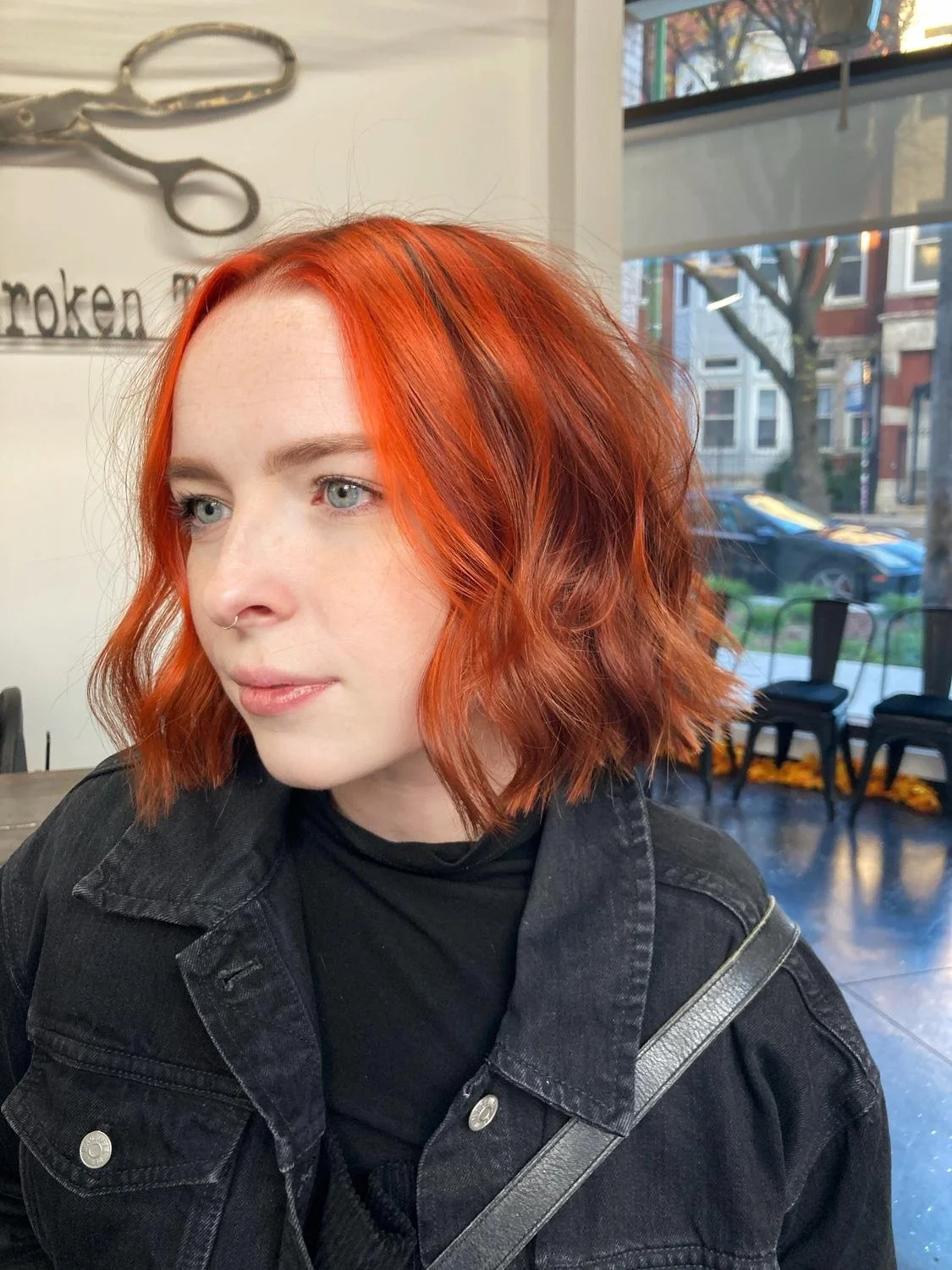 A woman with short, wavy red hair, wearing a black jacket, sitting inside a café near a window.
