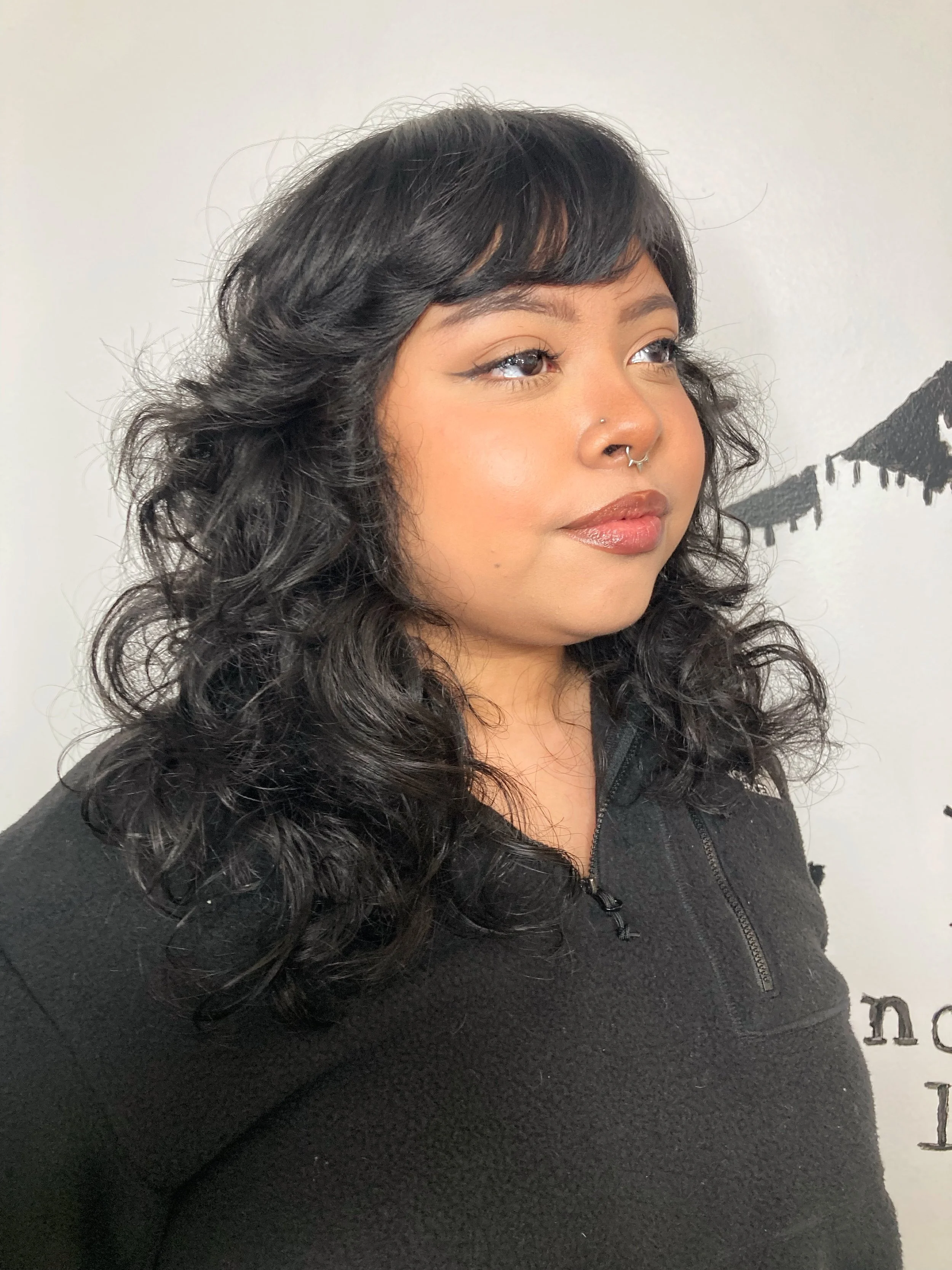 A woman with curly black hair, wearing a black zip-up top, looking to the side with neutral facial expression, in front of a white wall with black writing and designs.