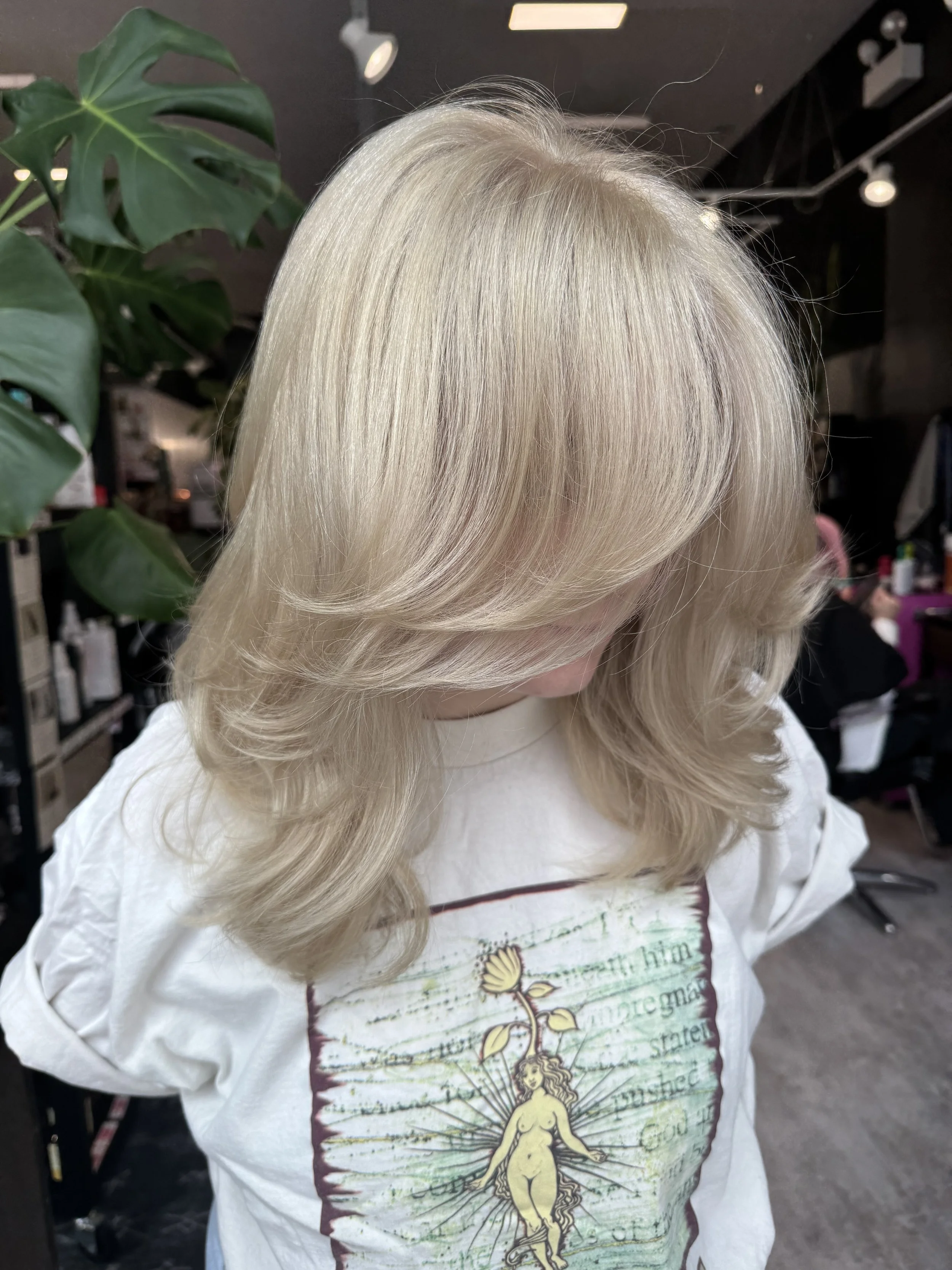 Blonde woman with styled, layered hair in a salon, wearing a white shirt with a graphic of a nude woman surrounded by text and a floral motif.