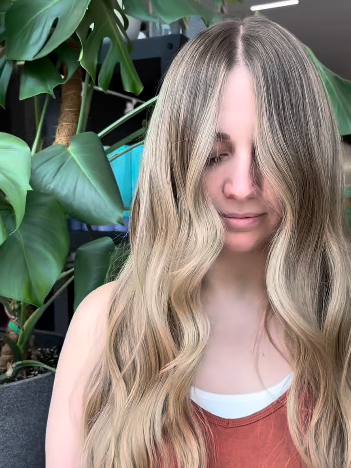 A woman with long wavy blonde hair and light skin, looking downward with her face partially obscured by her hair, standing in front of large green tropical leaves.