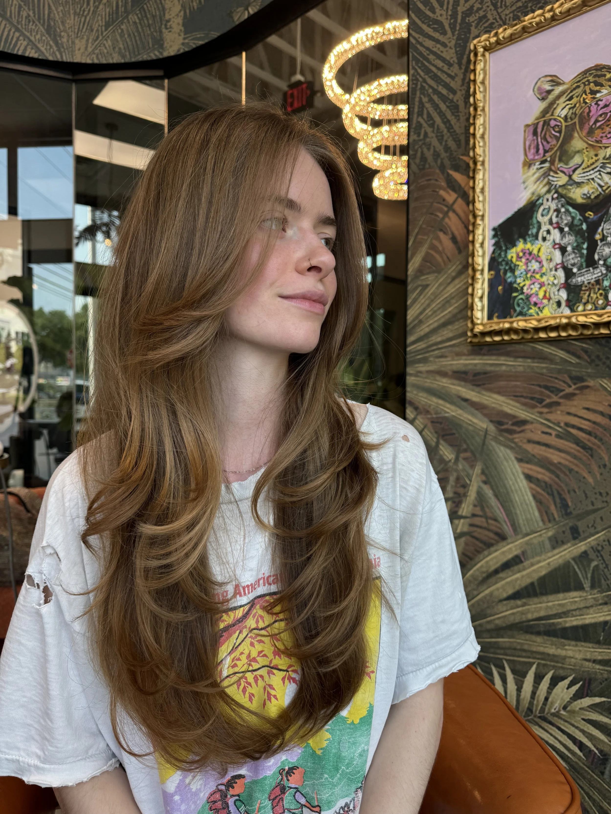 A young woman with long, wavy red hair and fair skin is sitting in a coffee shop, looking to her left. She is wearing a white T-shirt with colorful graphics and has a small nose piercing. Behind her, there is a framed painting of a tiger with glasses and jewelry, tropical-themed wallpaper, and modern lighting fixtures.