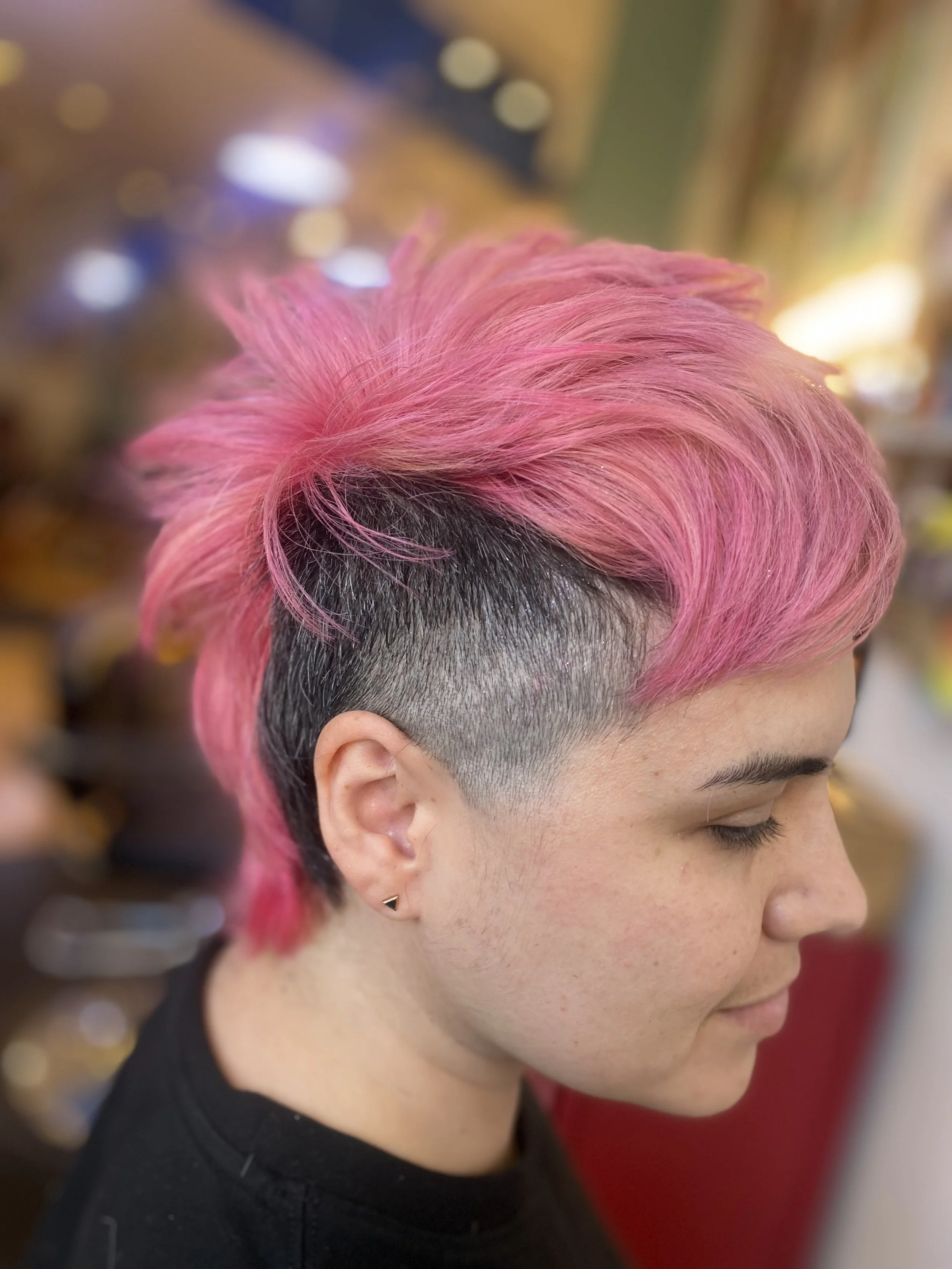 Side profile of a woman with short pink dyed hair, shaved on the sides, wearing small earrings.