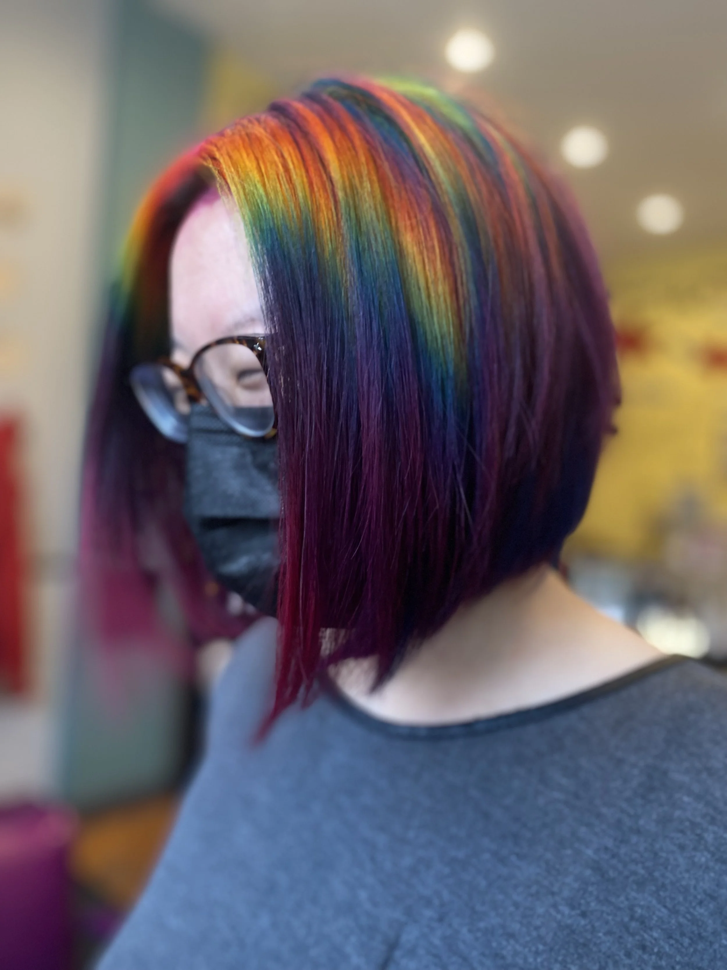 Woman with shoulder-length hair dyed in rainbow colors, wearing glasses and a black face mask.