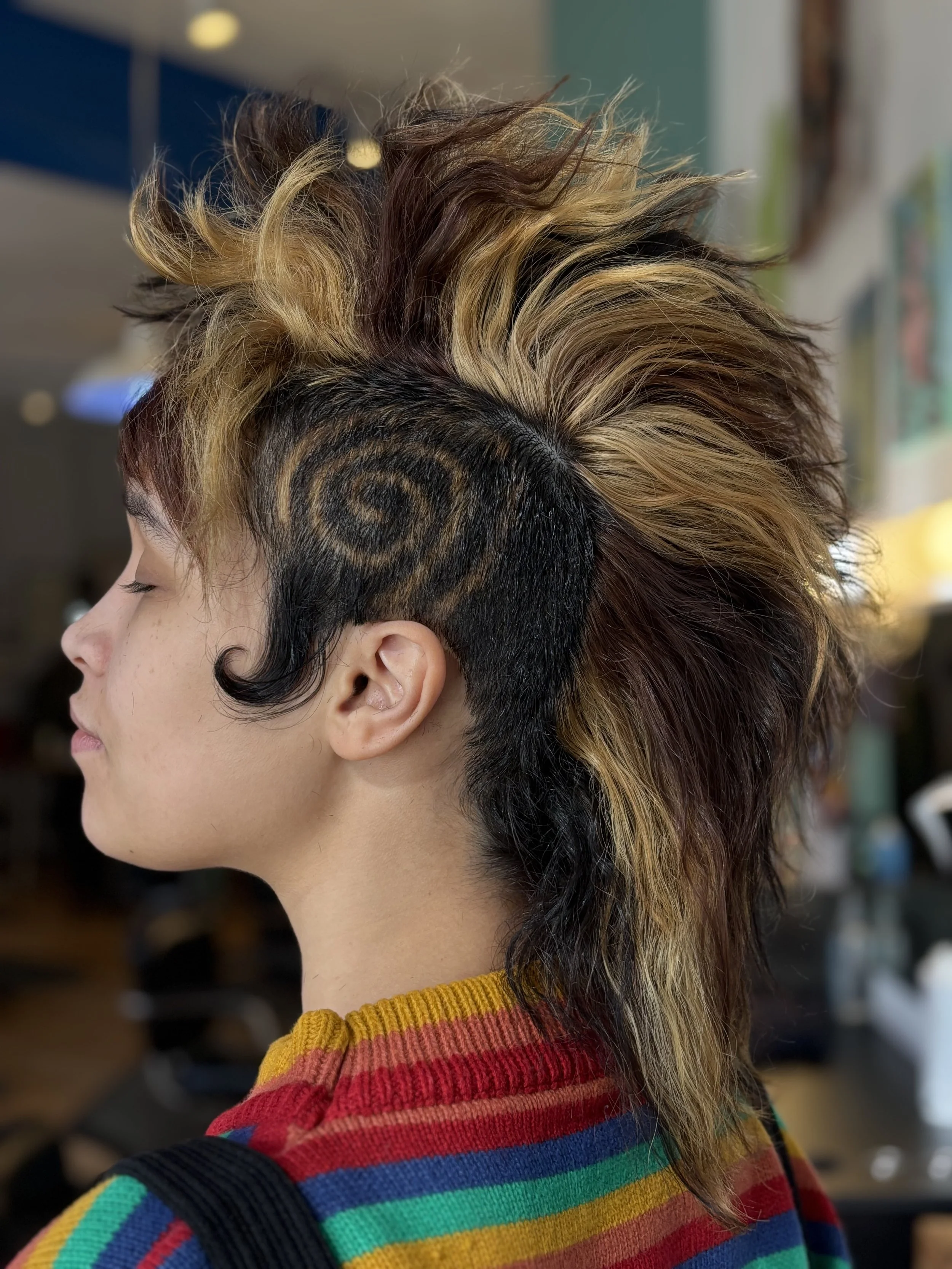 Side profile of a person with a colorful rainbow striped sweater and a punk hairstyle featuring a spiral pattern shaved into the side and voluminous, light brown and blonde hair on top.