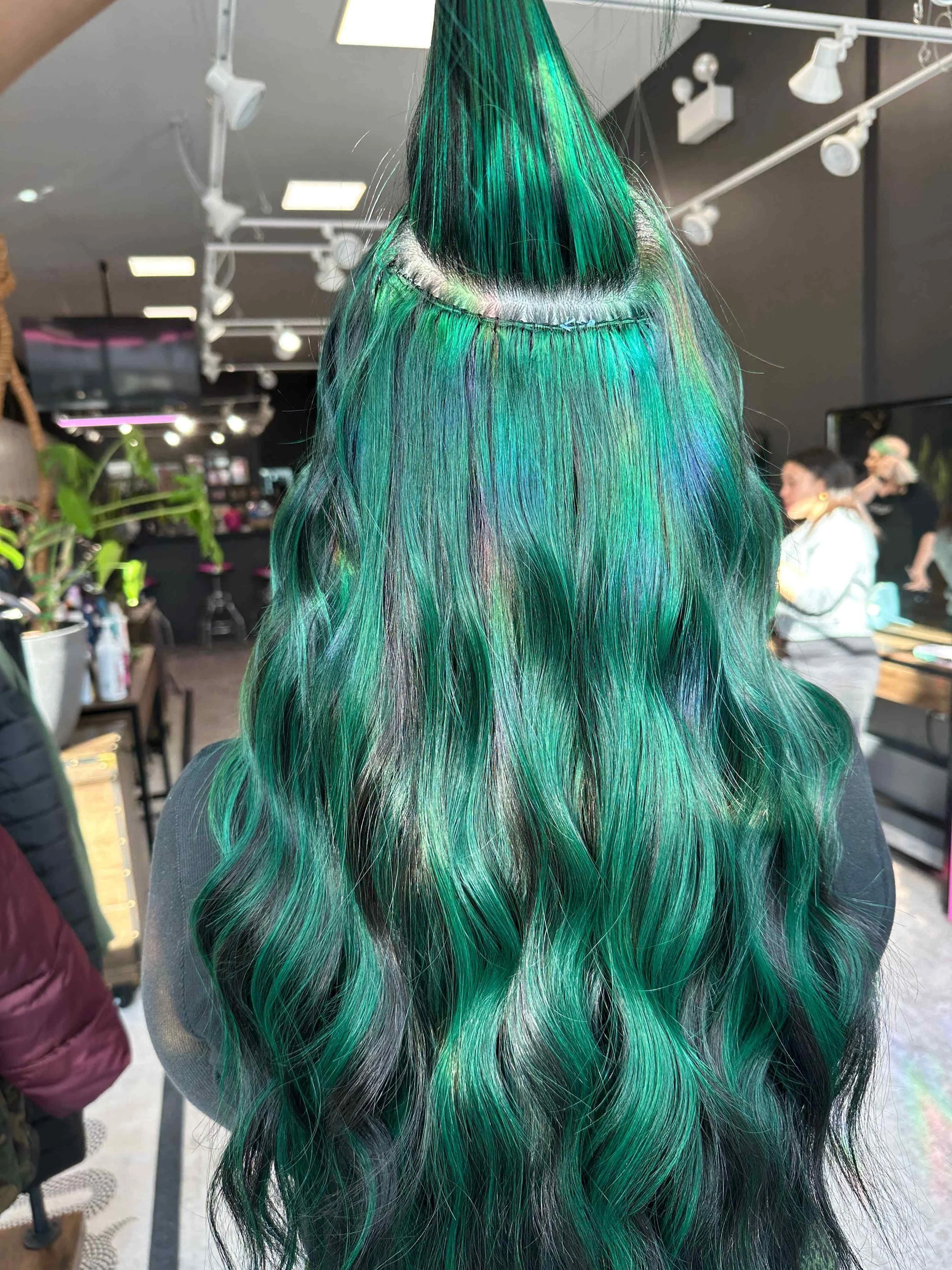 Back view of a woman's long, wavy hair dyed in vibrant shades of green with hints of blue, silver, and black in a salon setting.