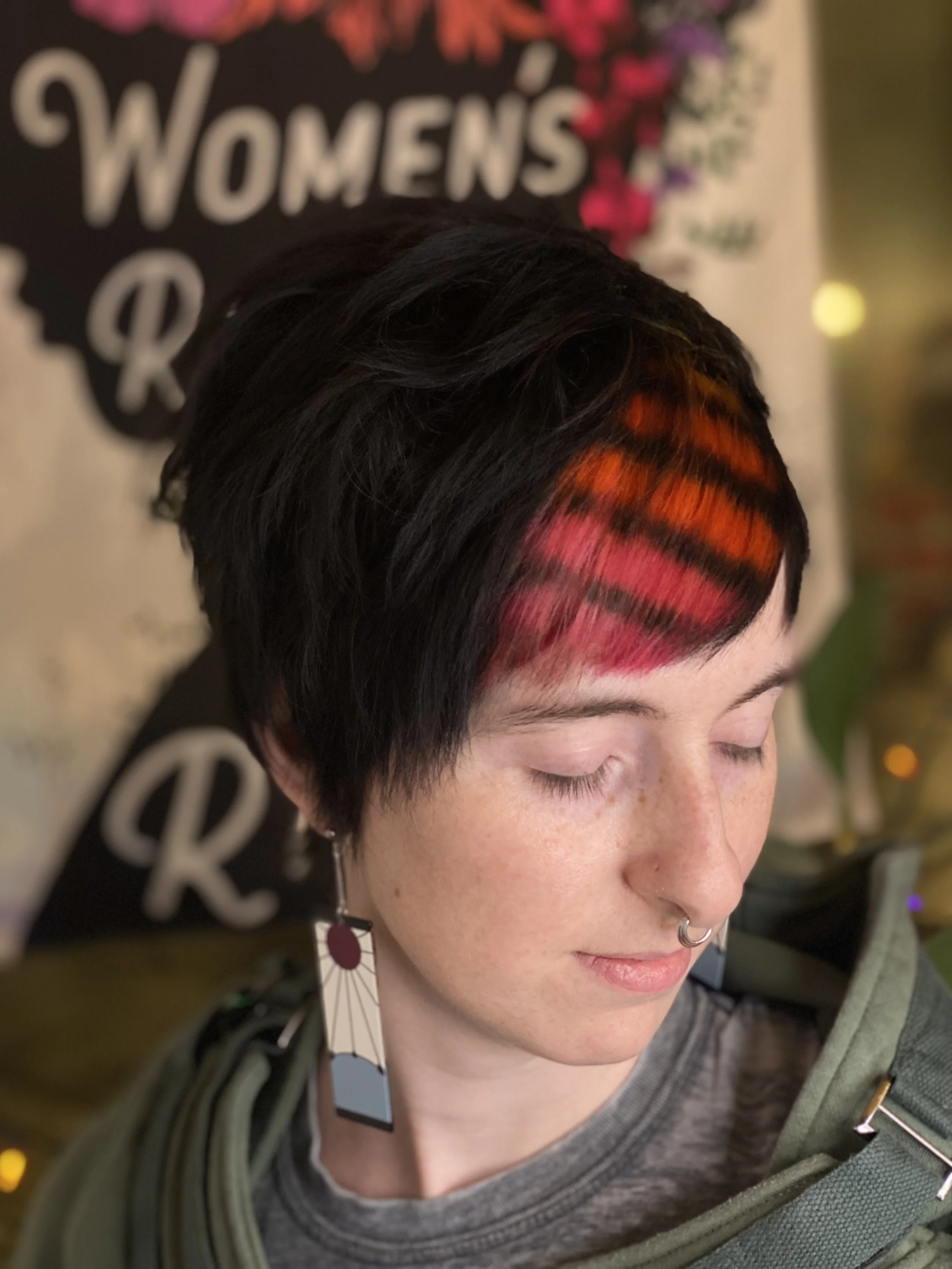 A person with short black hair featuring bright red and orange streaks, wearing a gray shirt, large geometric earrings, a septum piercing, and a green jacket. They are in front of a backdrop with the words 'Women's' and 'R' visible.