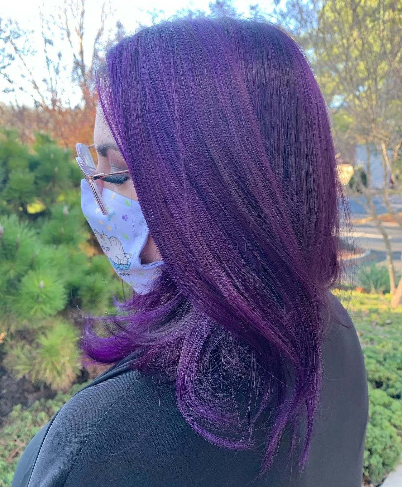 Profile of a woman with long, wavy, vibrant purple hair, wearing glasses and a face mask with a cartoon character, standing outdoors with trees and greenery in the background.