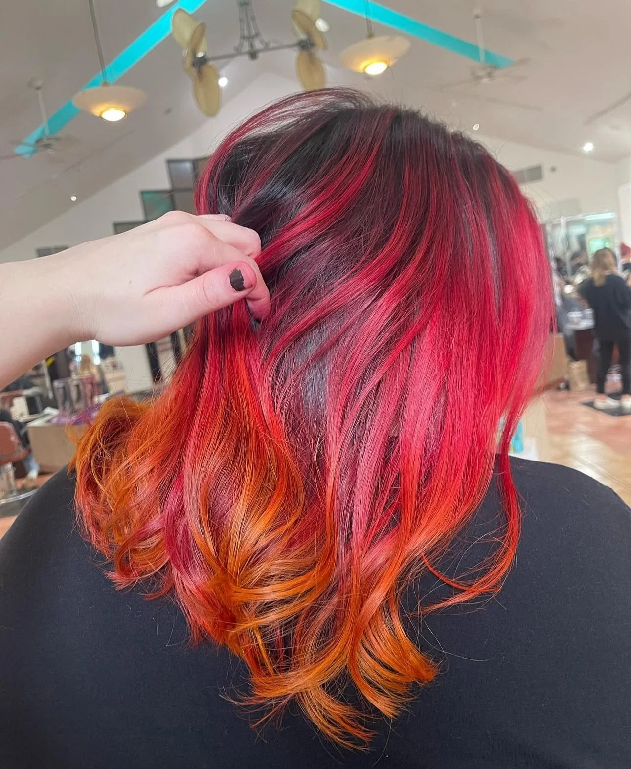 Back of a woman's head with vibrant red and orange ombre hair, styled in loose curls, inside a salon with other customers and hair products visible in the background.