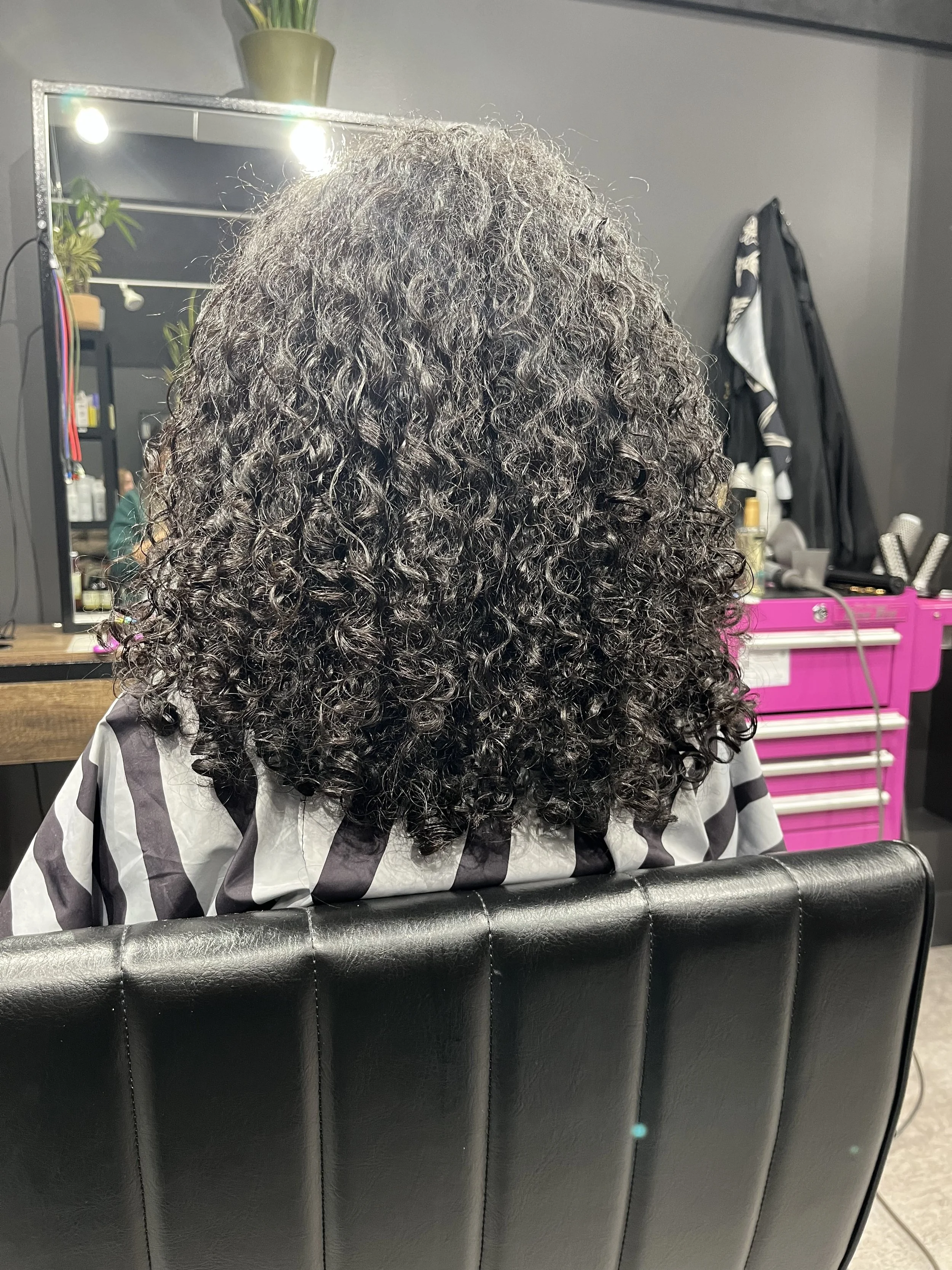 Back of a person with shoulder-length, curly black hair sitting in a salon chair, facing a mirror in a hair salon.
