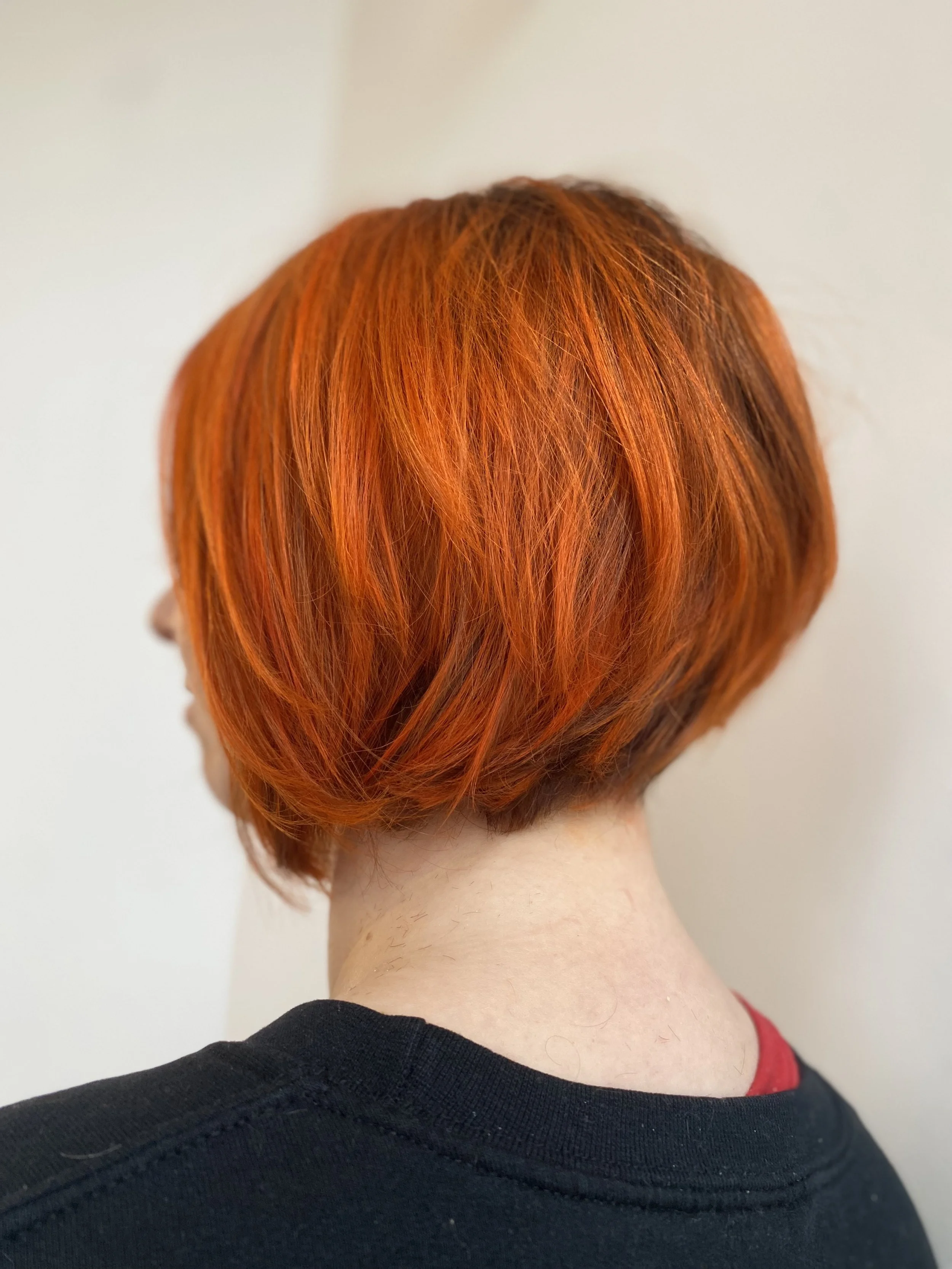 Back of a person's head with short, layered, bright orange hair, wearing a black shirt.