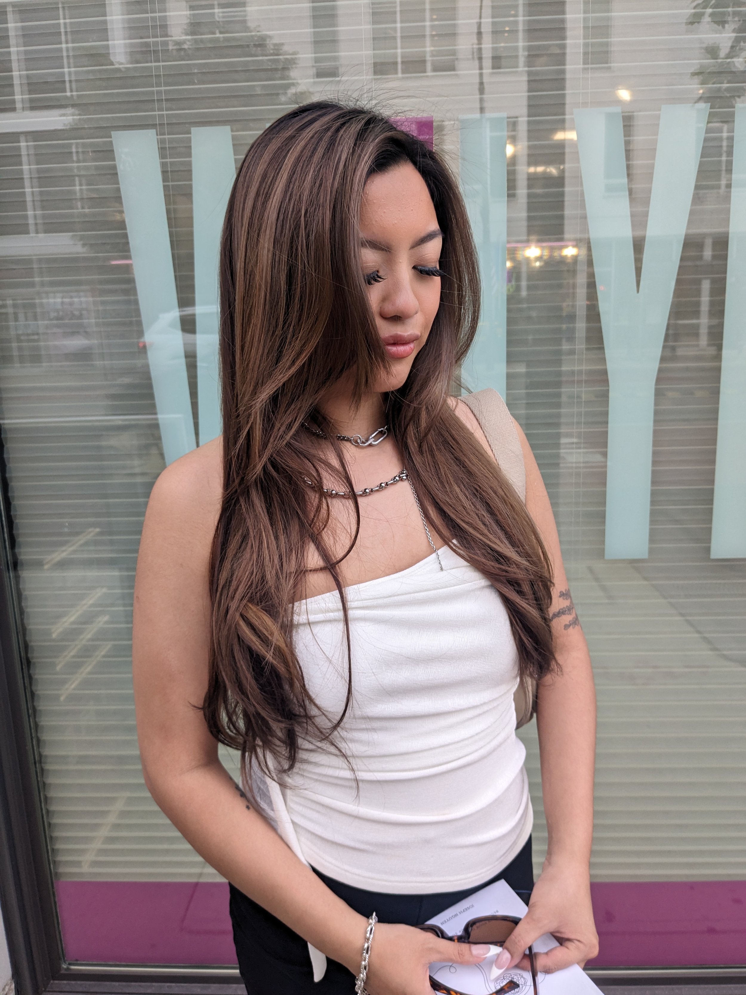 A young woman with long, wavy brown hair standing outside, wearing a white sleeveless top and silver jewelry. She is looking down and holding some papers in her hand, with city buildings and glass windows in the background.