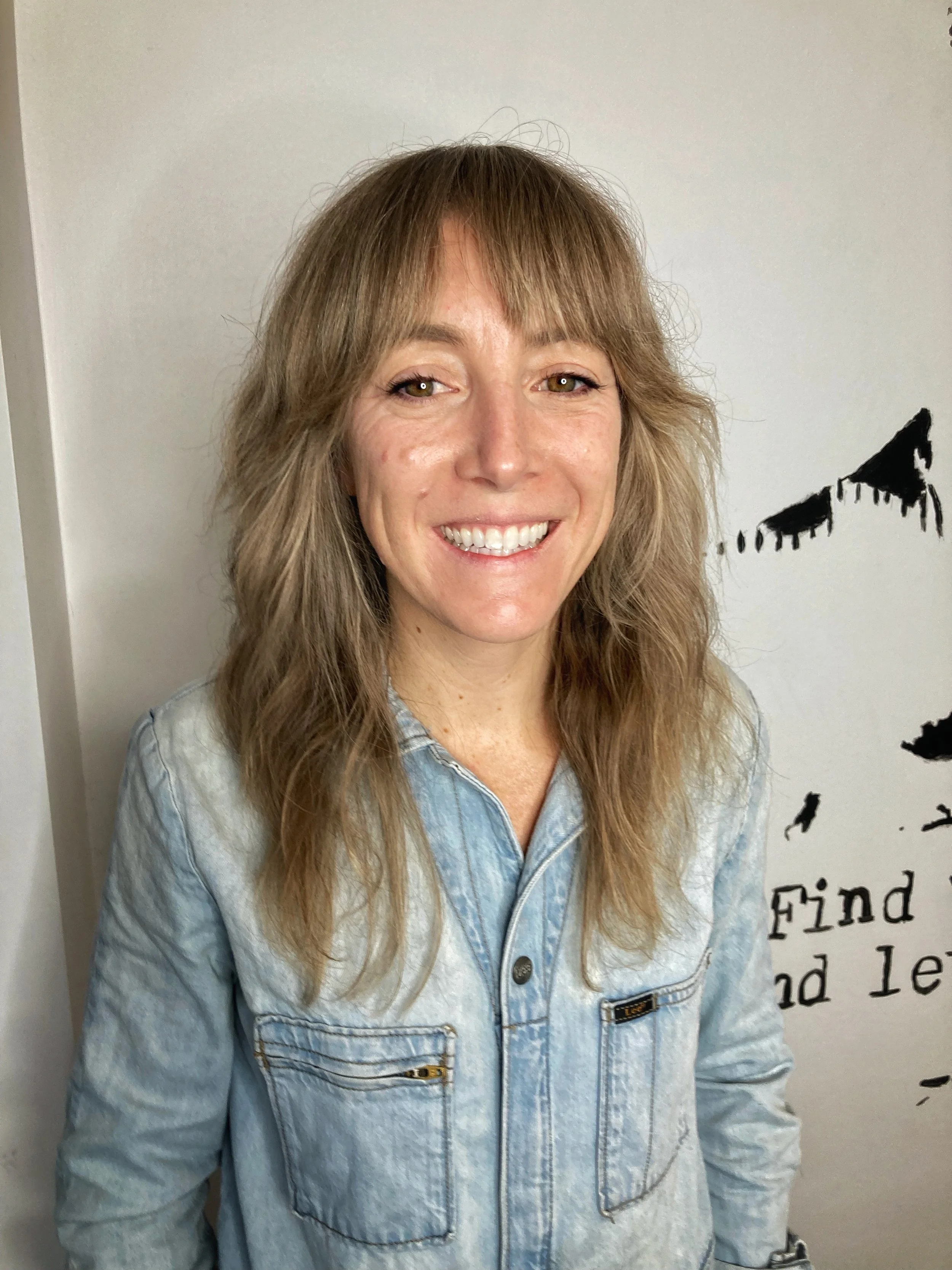 A woman with light brown hair smiling, wearing a light blue denim jacket, standing in front of a white wall with black painted letters and shapes.