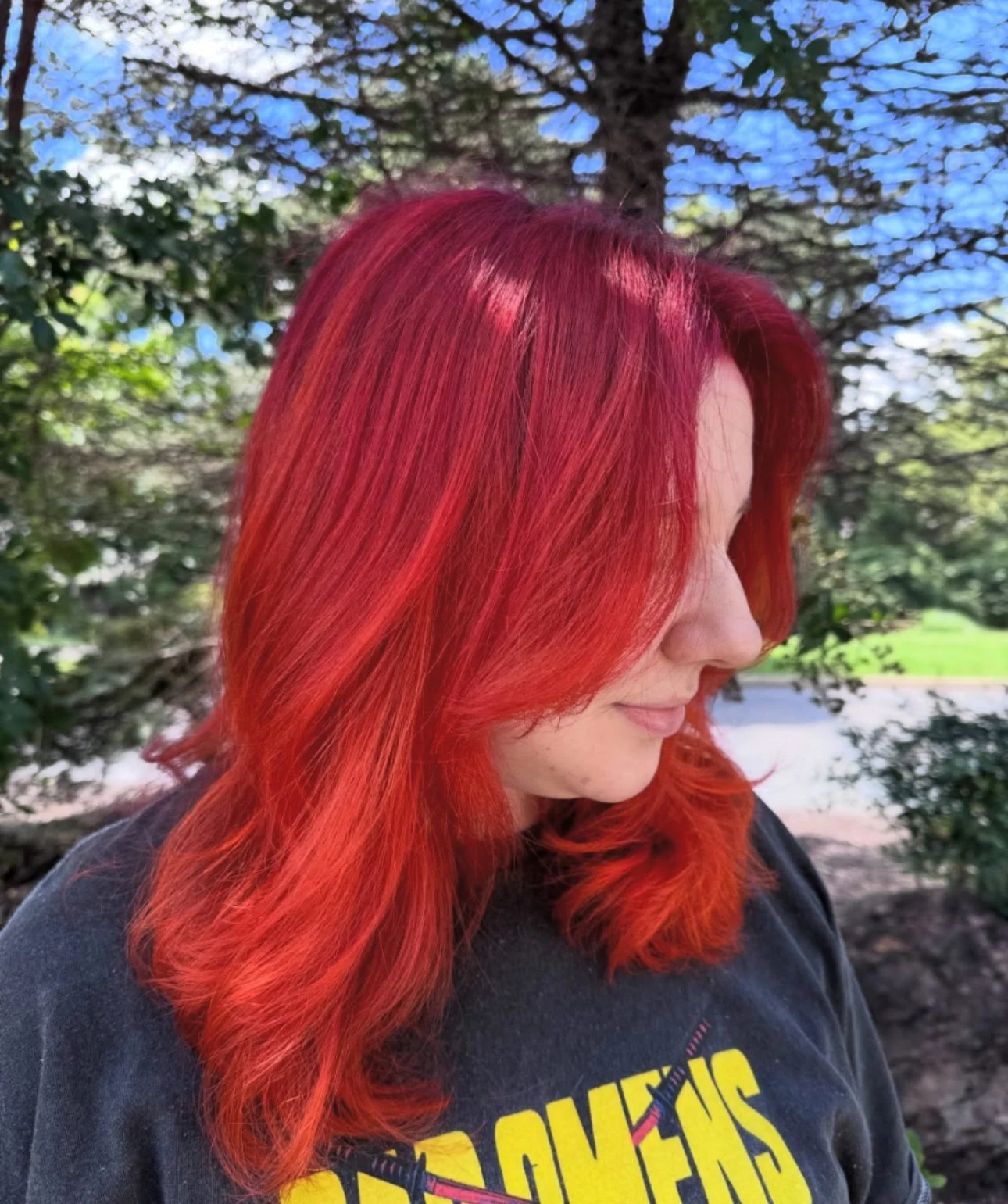 A woman with vibrant red hair standing outdoors near trees, wearing a black shirt with yellow text.