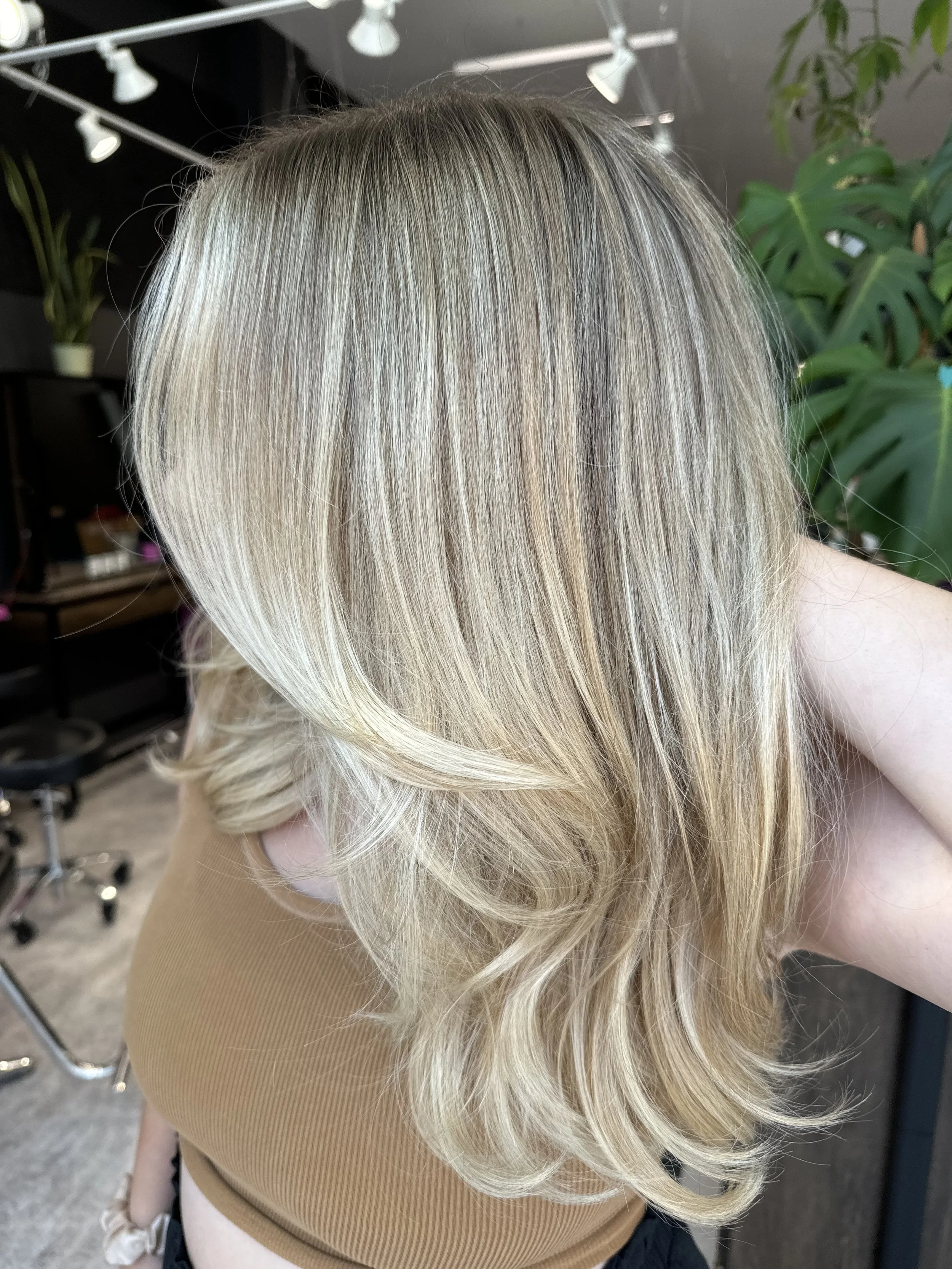 Blonde hair styled in loose waves with darker roots, sitting in a salon with plants and modern decor.