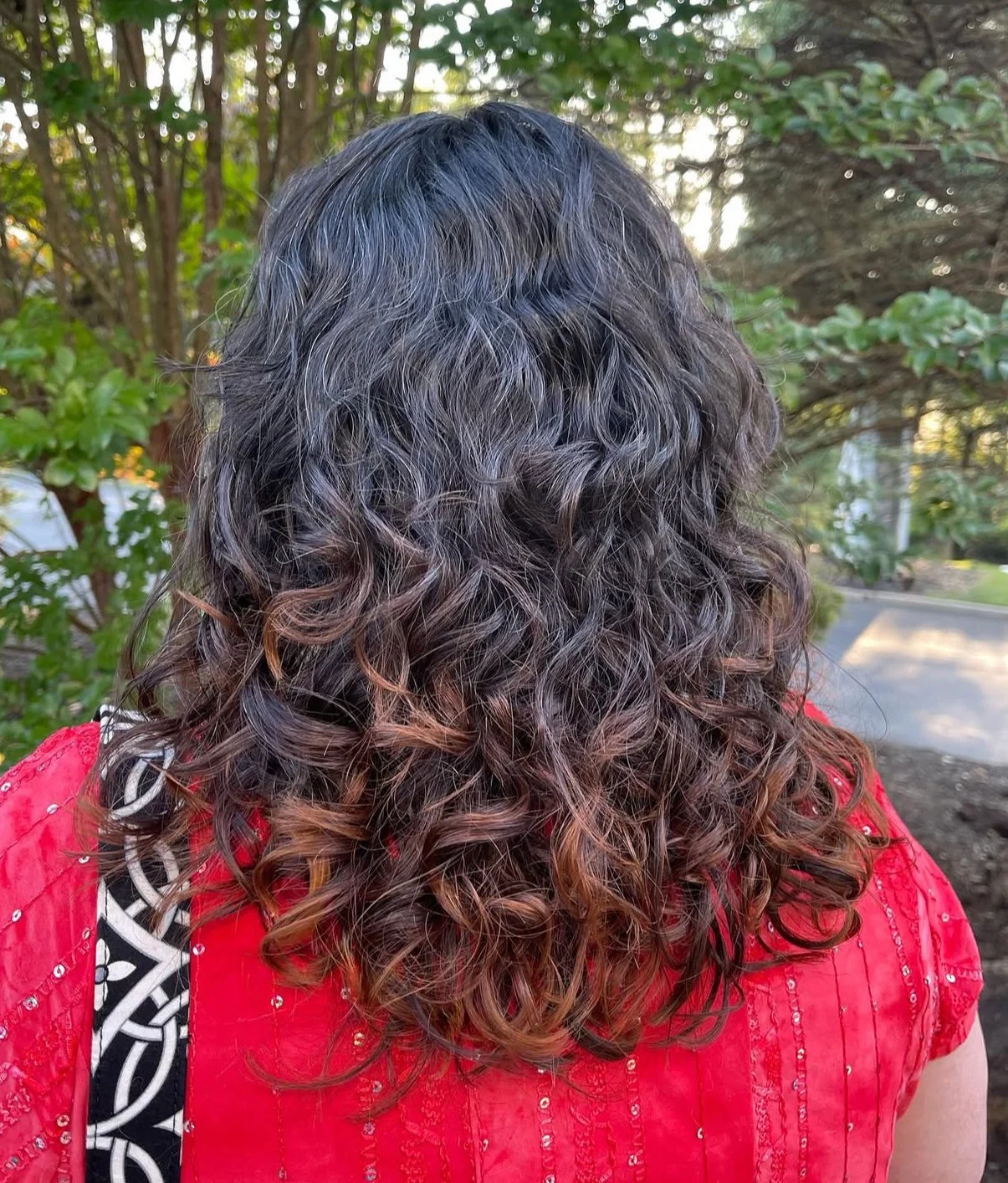 Back view of a woman with long, curly hair having dark brown to light brown ombre color, wearing a red top with black and white patterned strap, outdoors with trees and shrubs.
