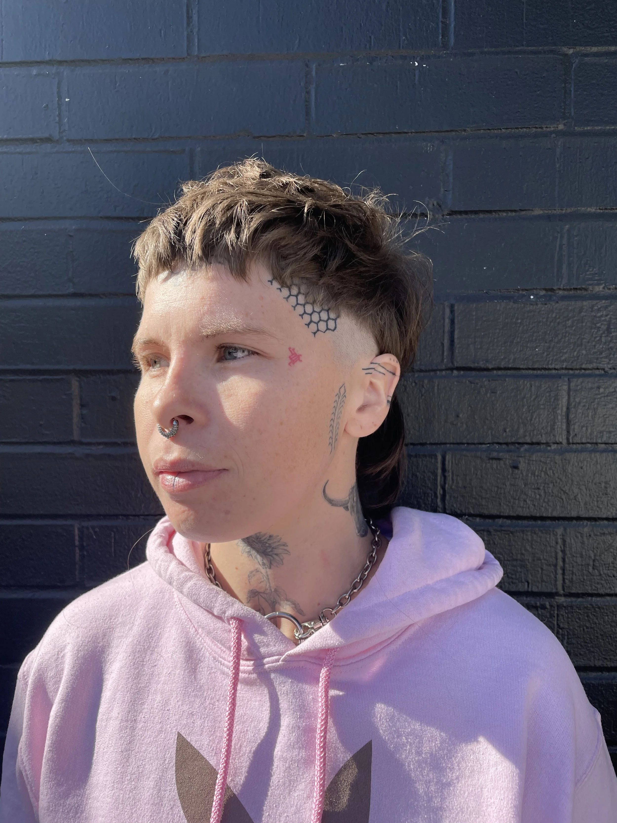 A person with short brown hair with a shaved side and multiple tattoos, wearing a pink hoodie and a chunky silver chain necklace, standing against a black brick wall.