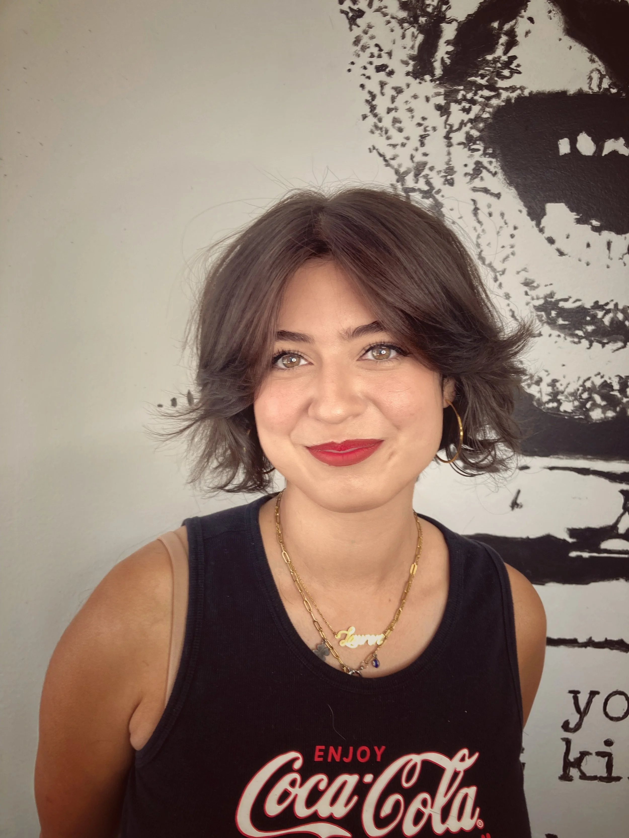 A young woman with short, wavy brown hair and gold hoop earrings, wearing red lipstick and a black Coca-Cola tank top, stands in front of a black and white abstract background with a partially visible face.