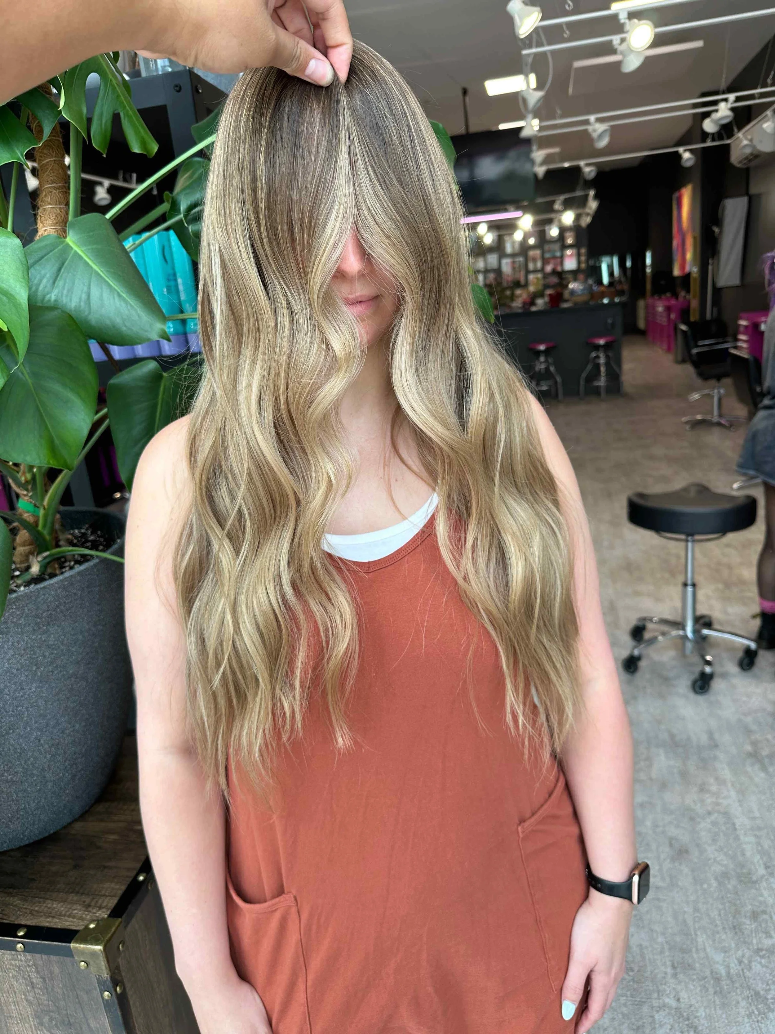 A woman with long wavy blonde hair wearing an orange sleeveless dress, sitting in a hair salon with her eyes covered by her hair, while a stylist holds a section of her hair.