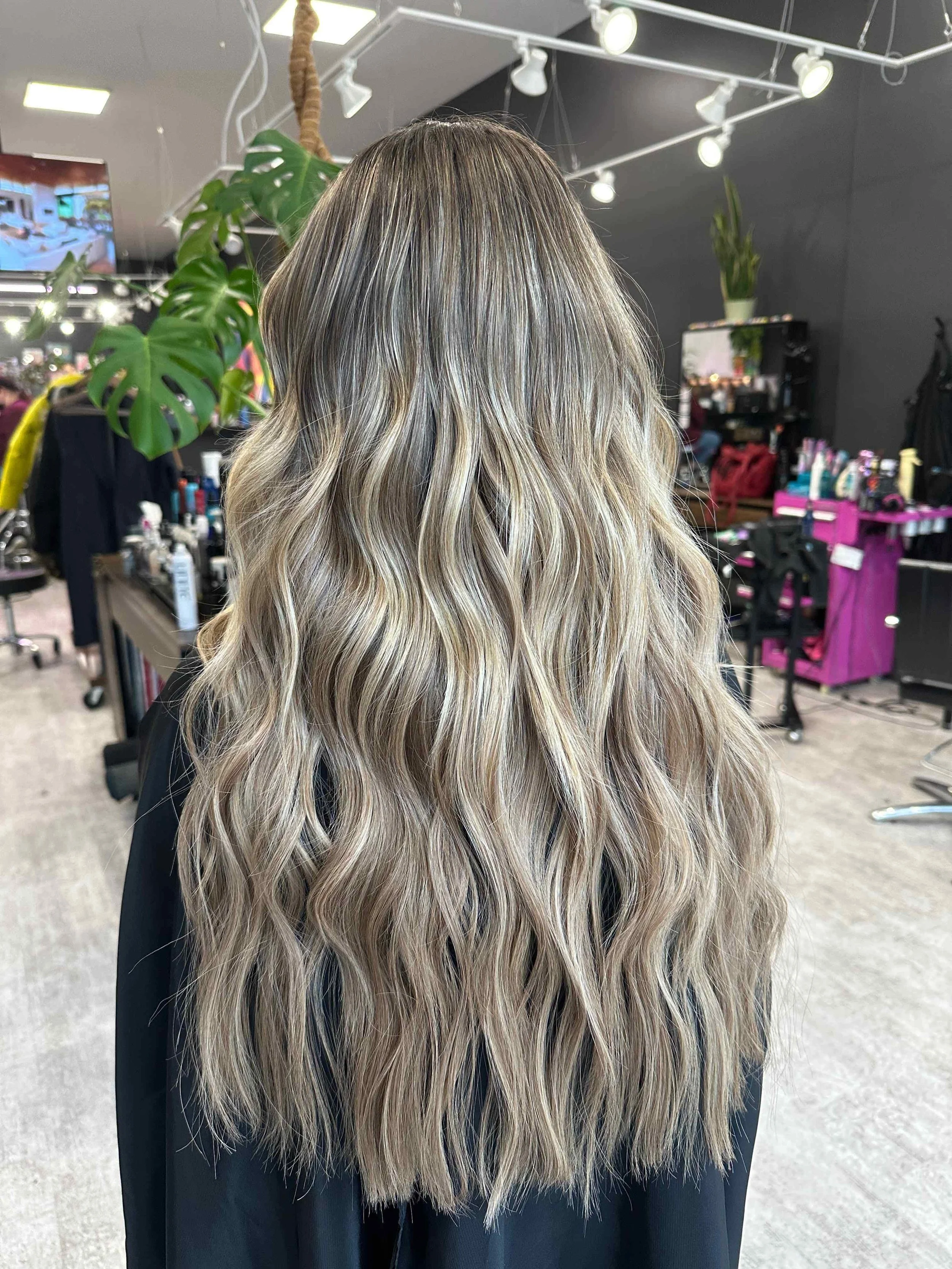 Back view of a woman with long, wavy blonde hair in a hair salon.