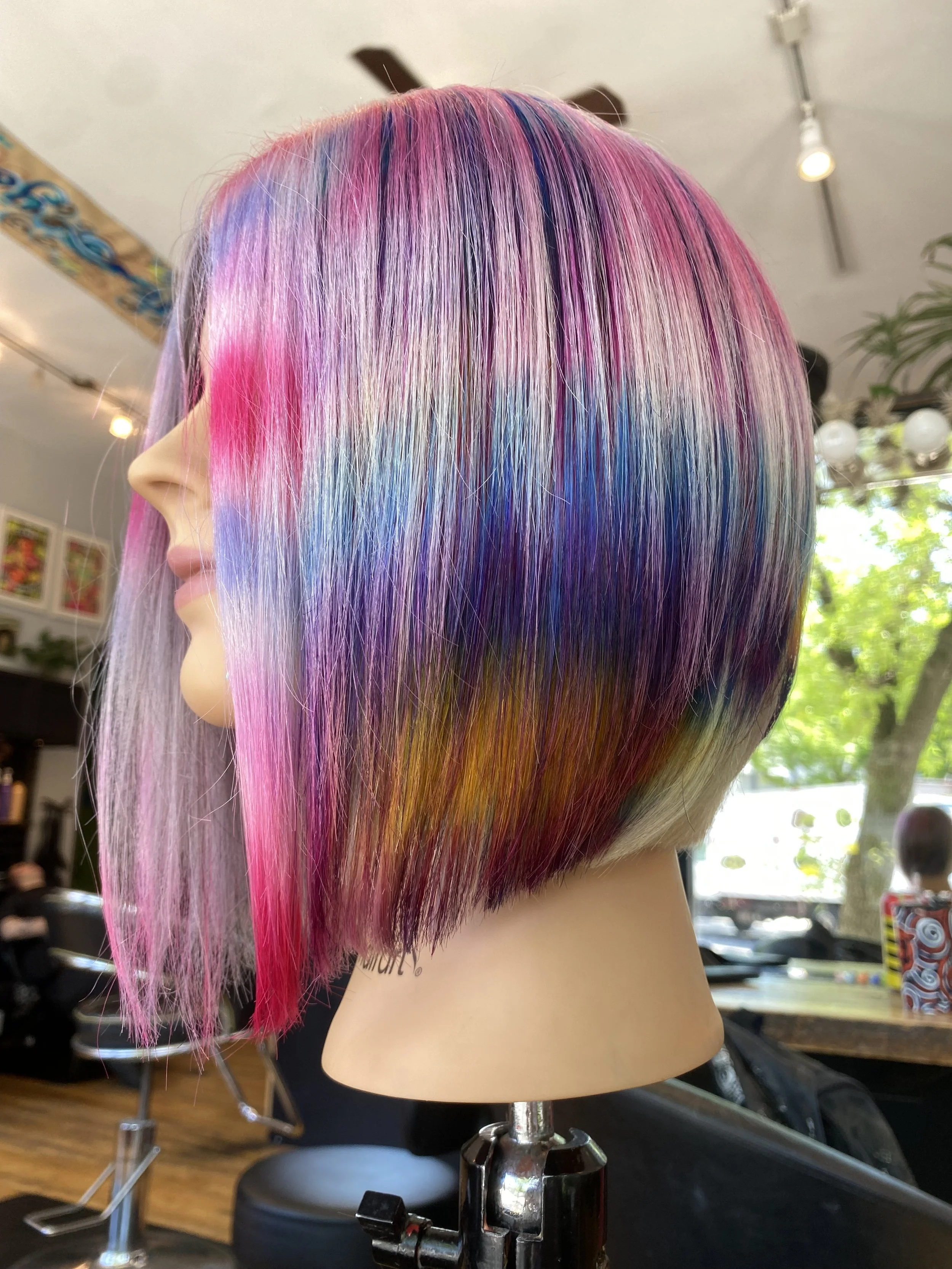 A mannequin with a multicolored, vibrant hairstyle featuring shades of pink, purple, blue, and blonde, styled in a sleek, straight bob cut inside a salon.