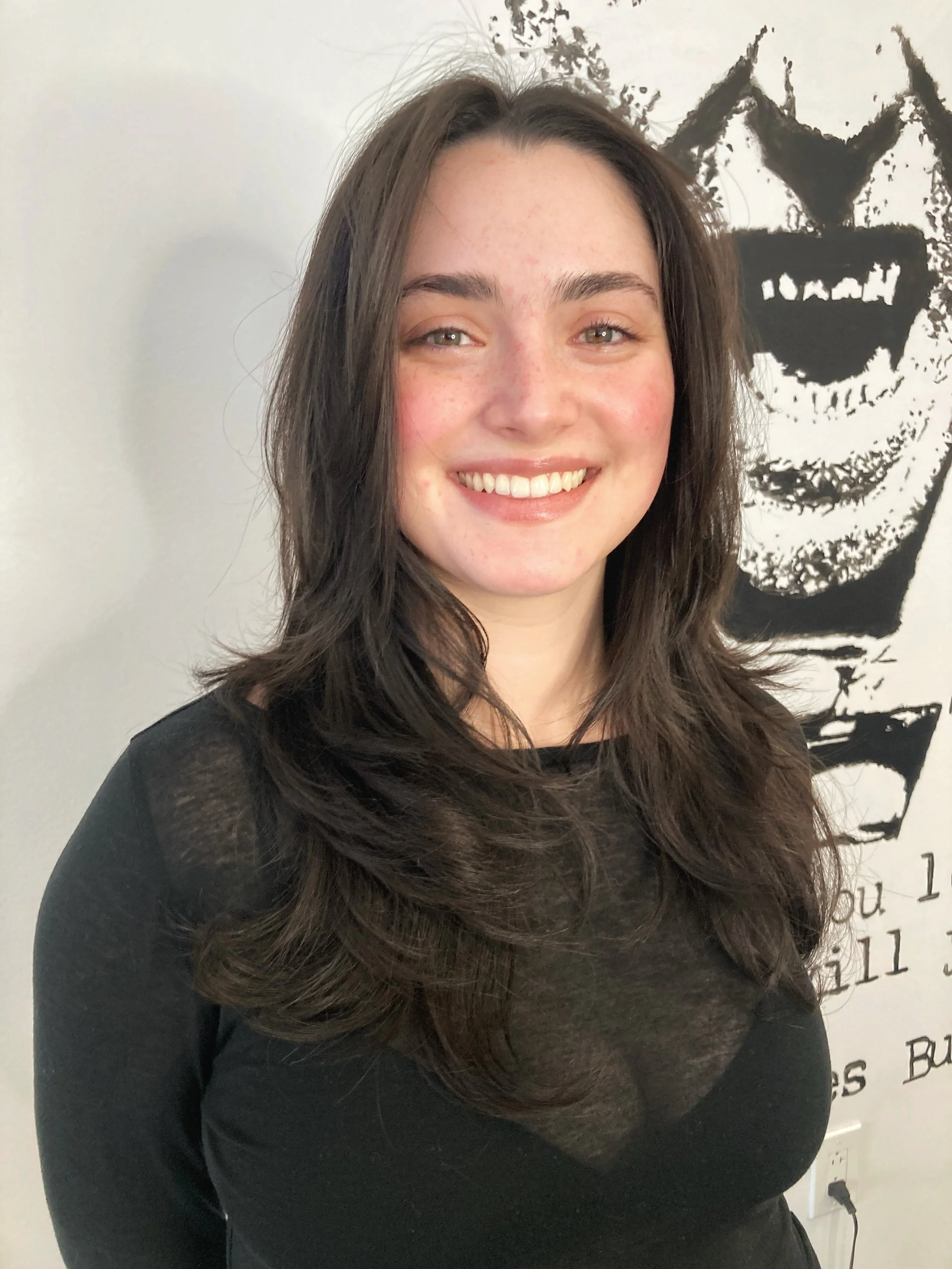 A young woman with long, wavy brown hair and light skin, smiling in front of black and white wall art of a face with a wide grin.