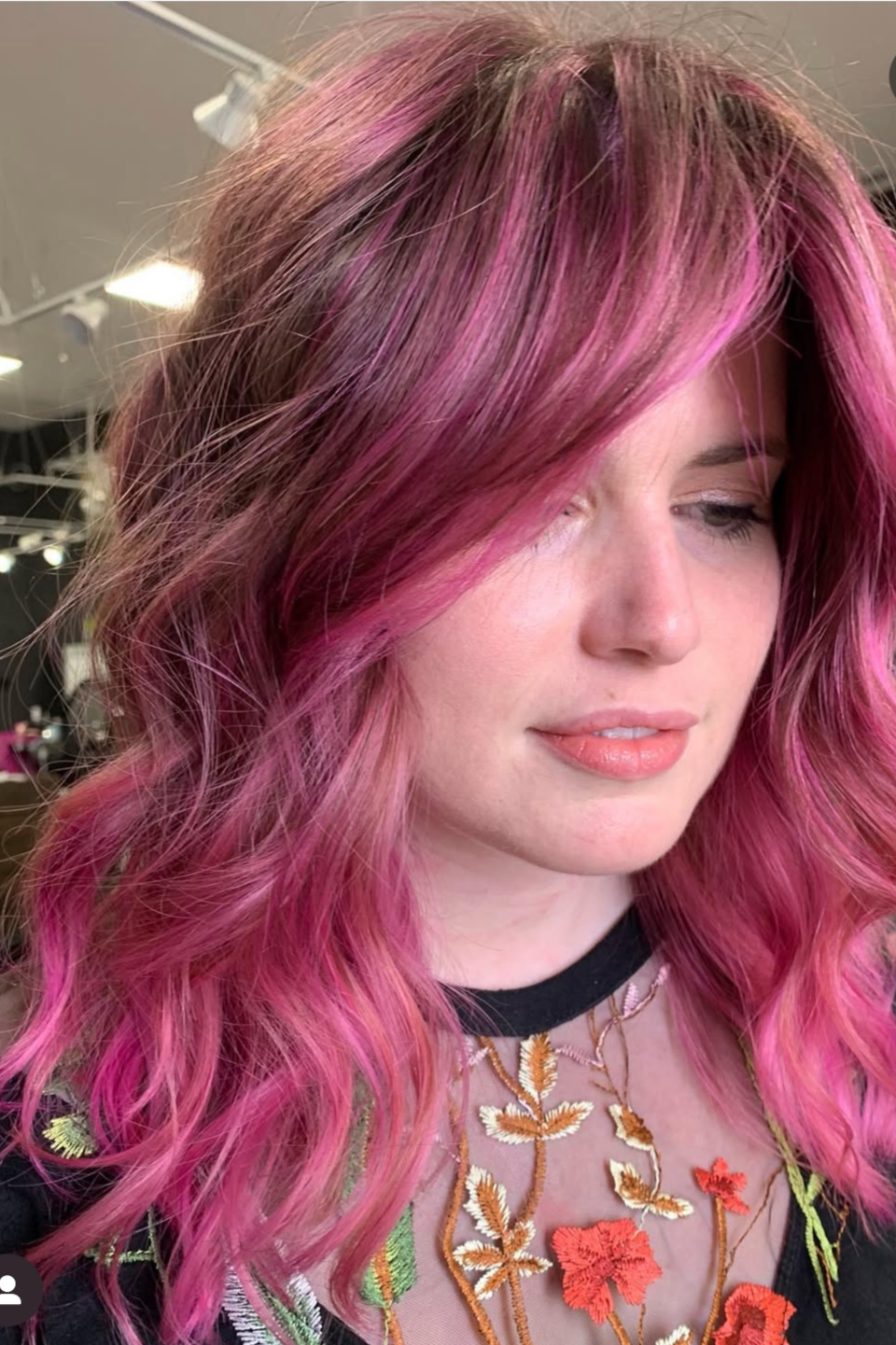 A woman with pink ombre hair, wearing a sheer top with floral embroidery and a black choker in a salon or studio setting.