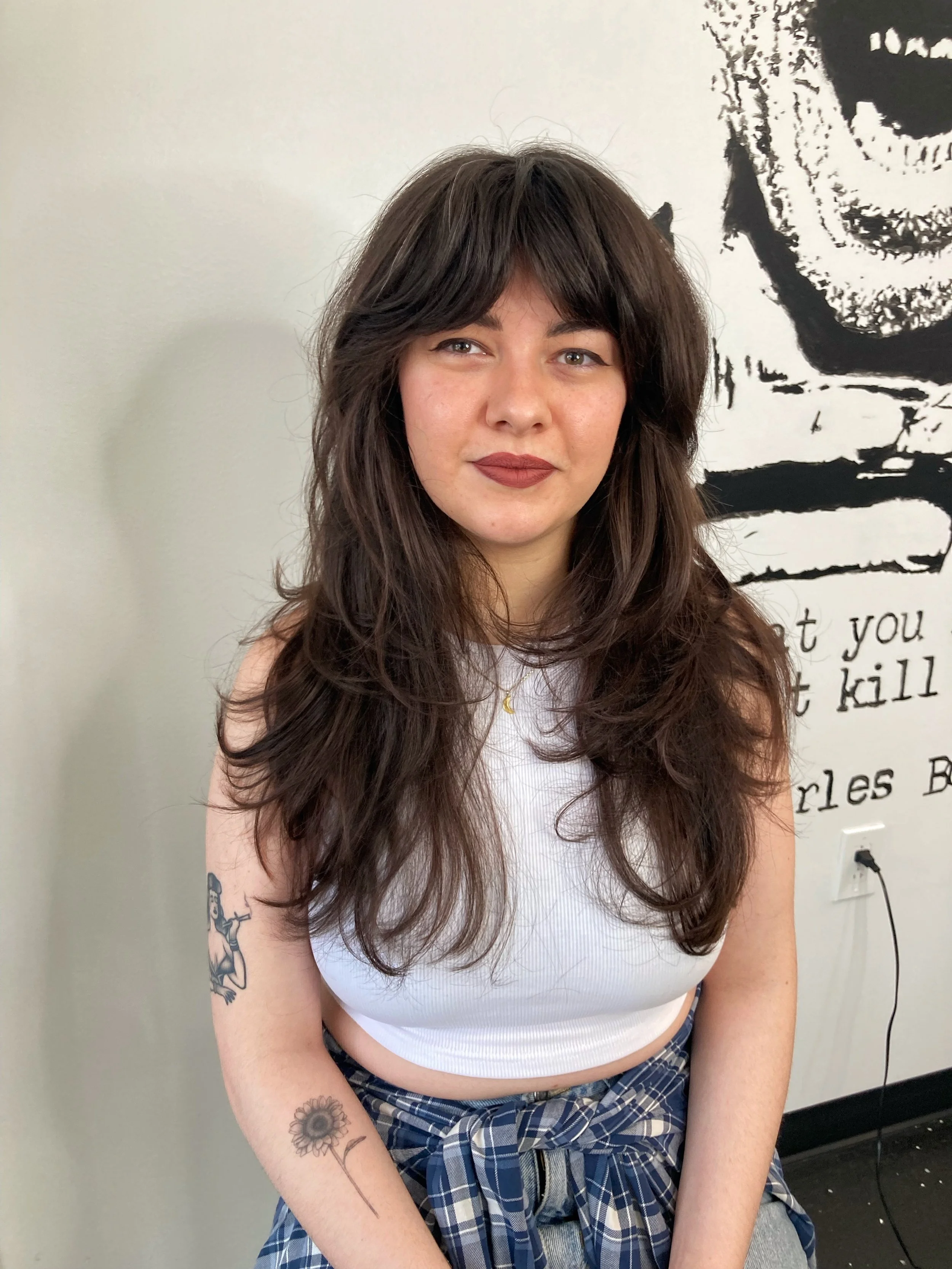 Young woman with long wavy brown hair, wearing a white crop top and plaid shirt tied at the waist, standing indoors in front of a black and white mural with text.