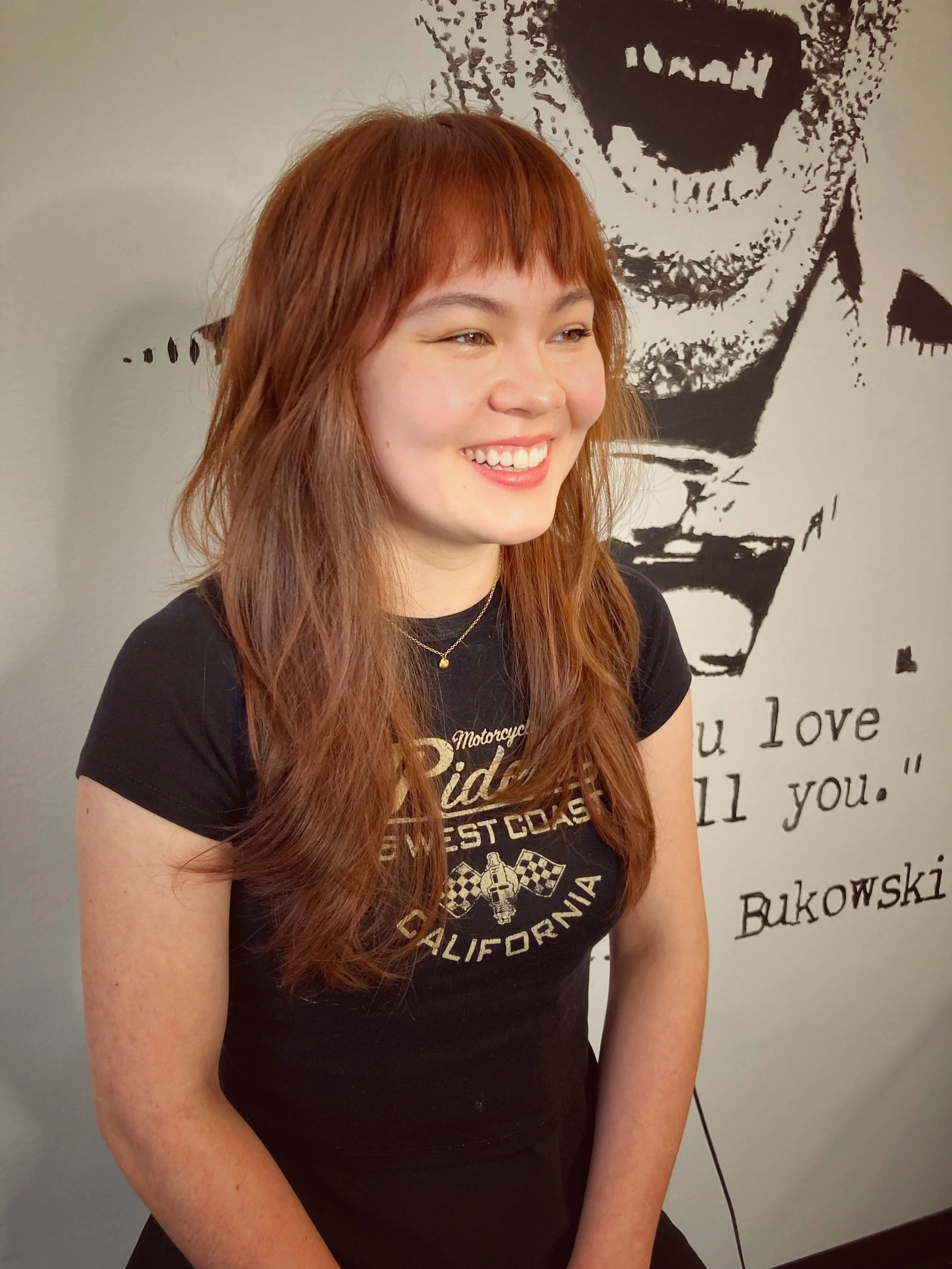 A young woman with long red hair smiling, standing in front of a black-and-white wall art of a face with a quote that says, 'I love you all you.'
