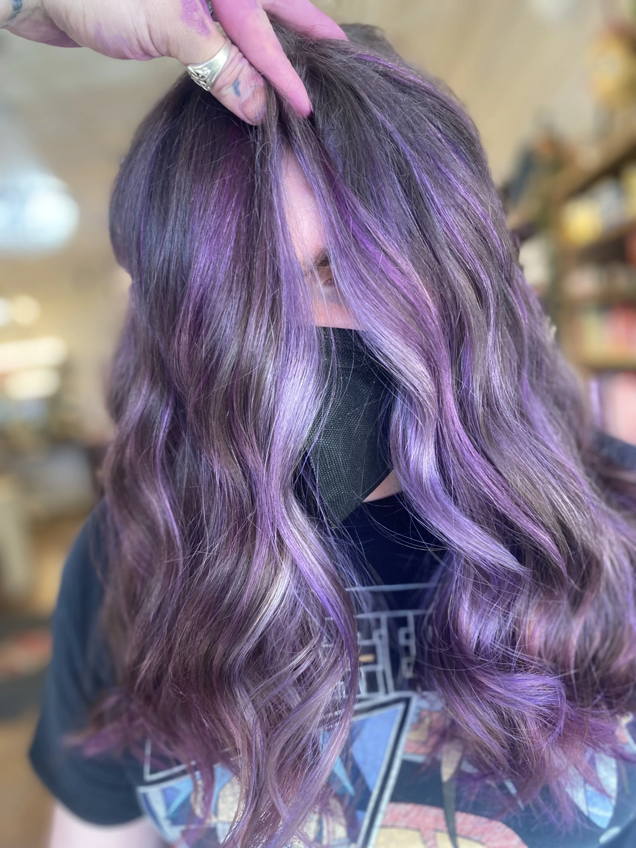 Person with long, wavy purple hair, wearing a black face mask and a dark graphic t-shirt, in a salon setting.