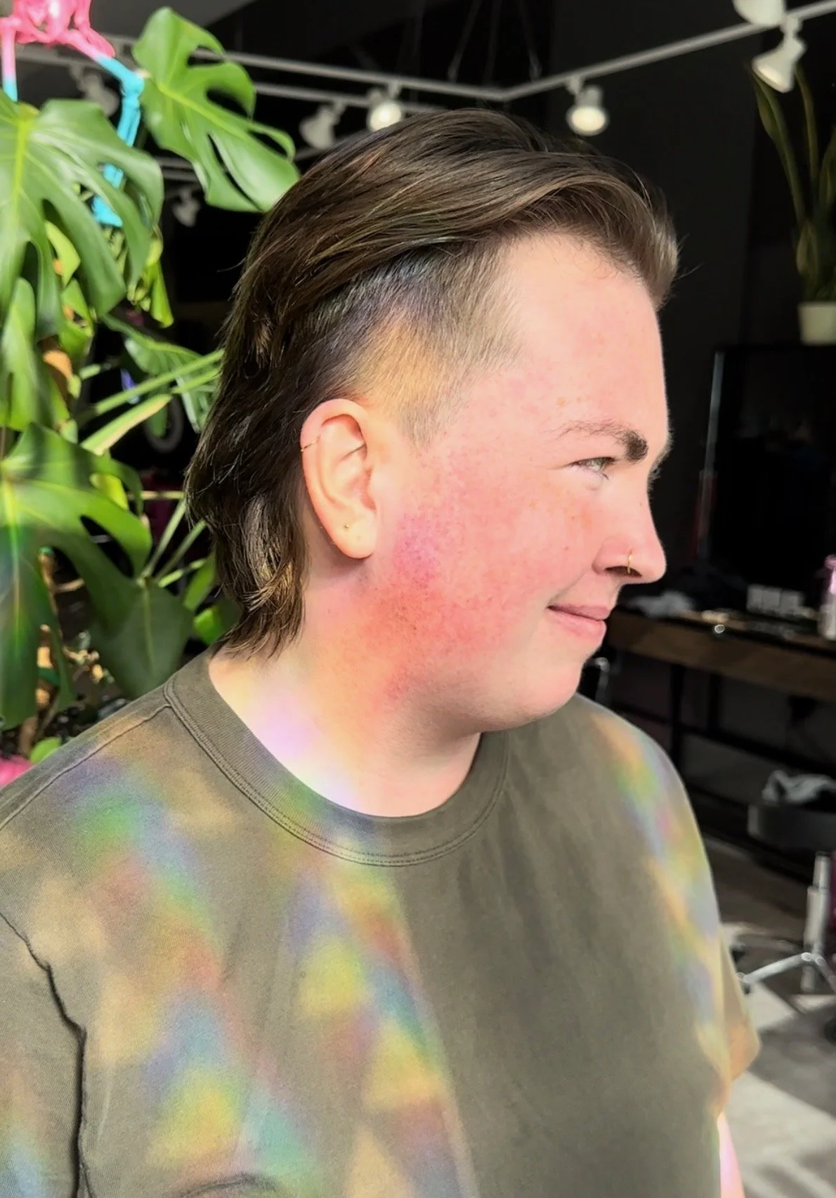Person with short side-swept brown hair, light skin, and a nose ring smiling in a room with green plants and colorful light reflections.