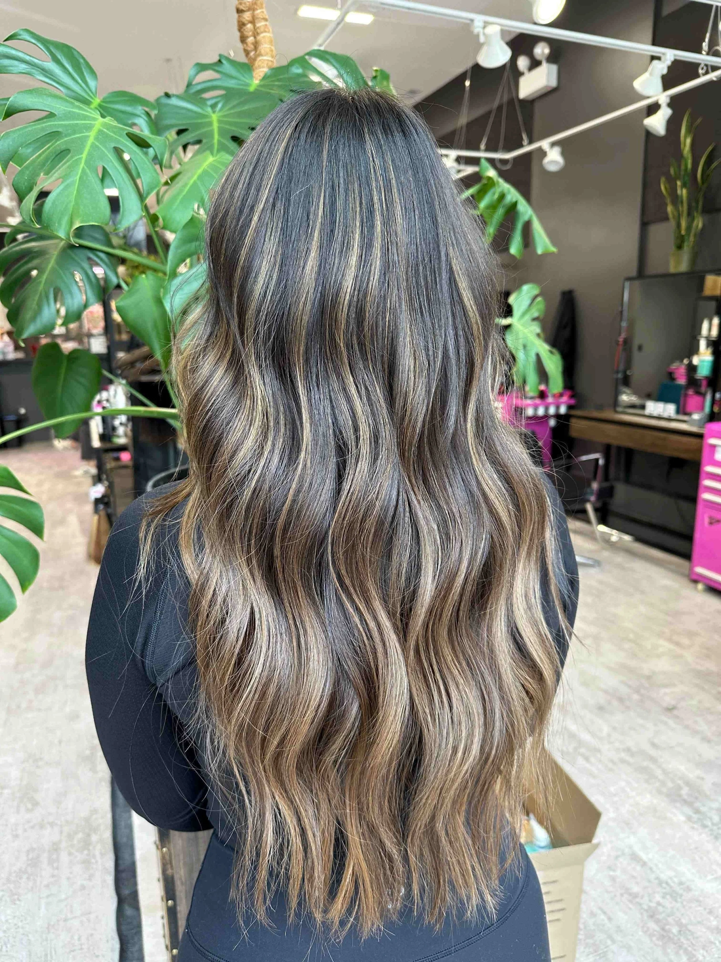 Back view of a woman with long, wavy hair featuring dark roots and blonde highlights, inside a salon with green plants and pink salon equipment.