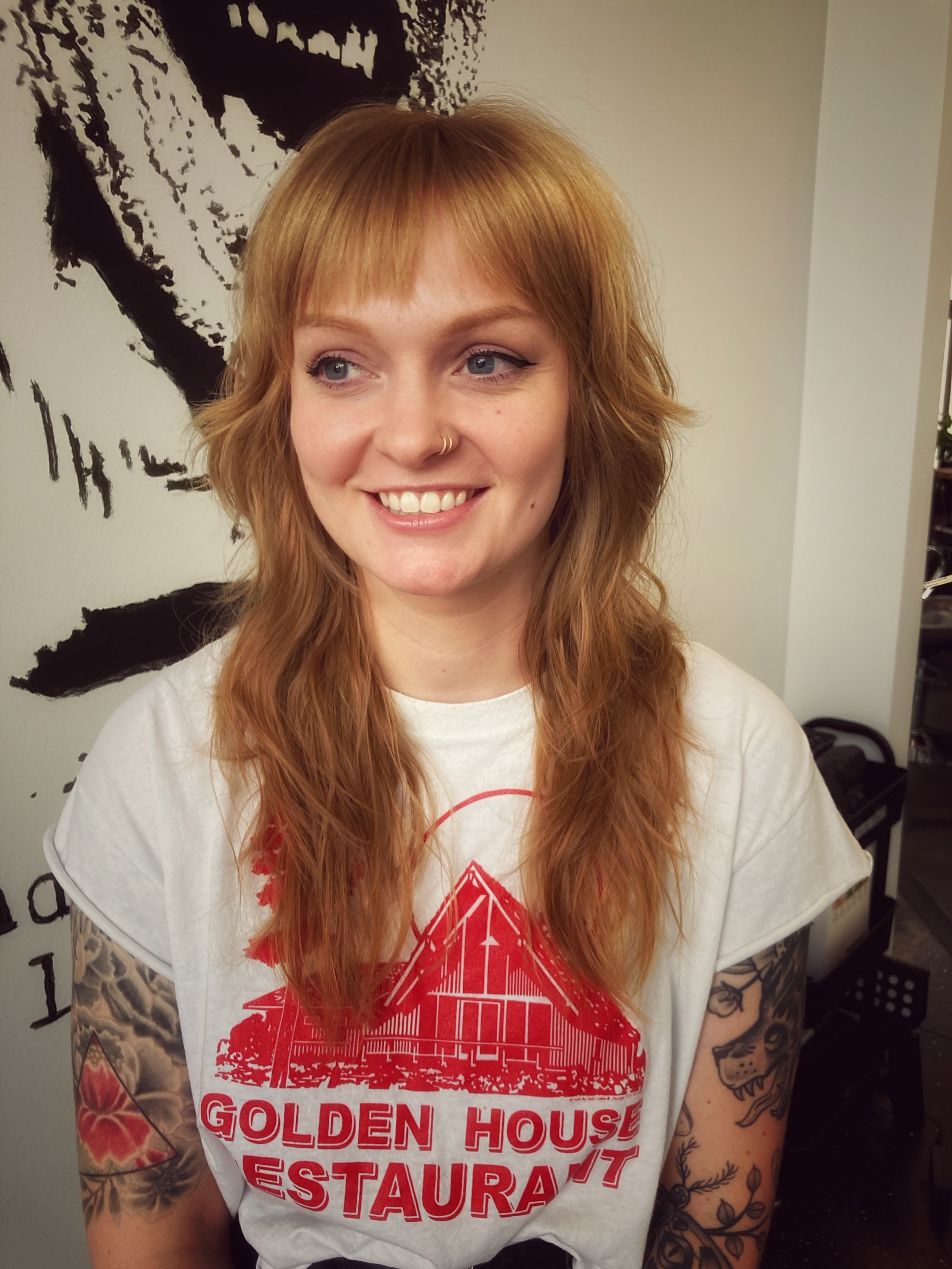 A woman with wavy red hair, light blue eyes, and tattoos on her right arm, smiling in front of a black and white artwork on the wall. She is wearing a white T-shirt with red print and a small nose piercing.