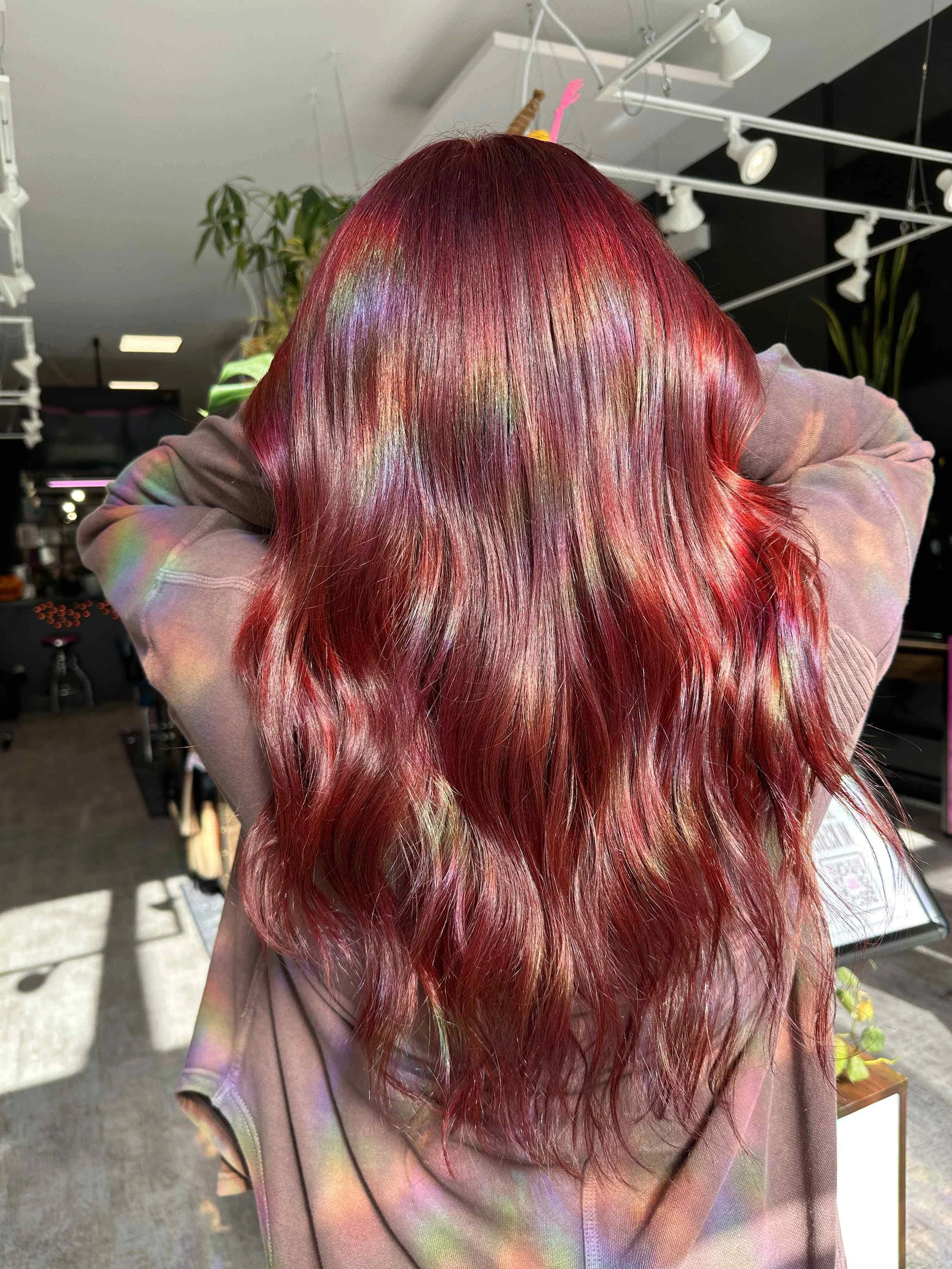 Person with long, wavy hair dyed in vibrant red and purple shades, shown from the back in an indoor setting.