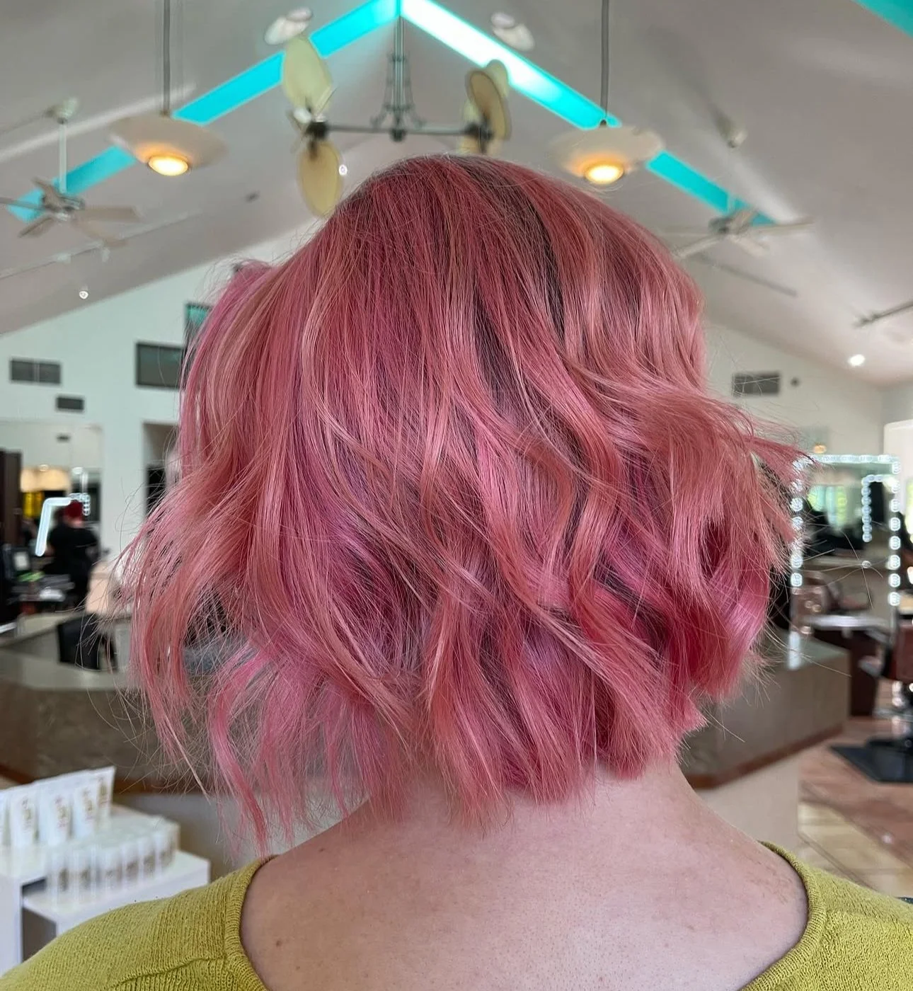 A person with short, wavy pink hair in a salon with ceiling fans and blue lighting.